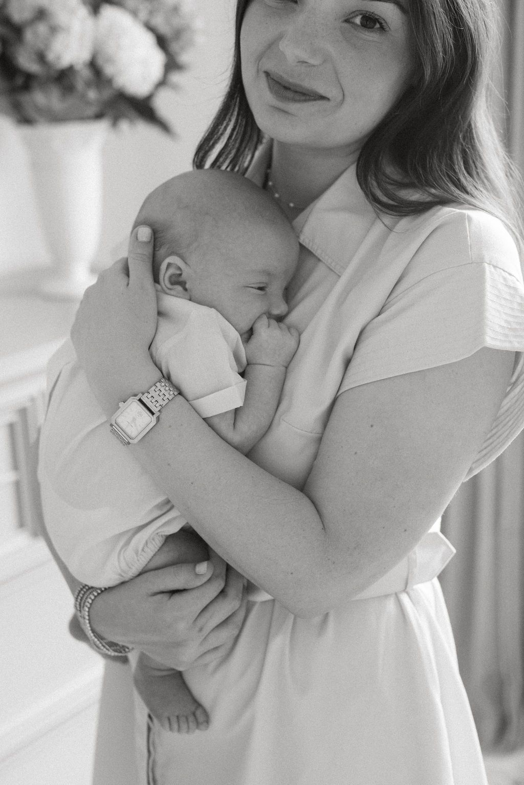 Black and white portrait of a mother holding her sleeping newborn baby near floral arrangements.