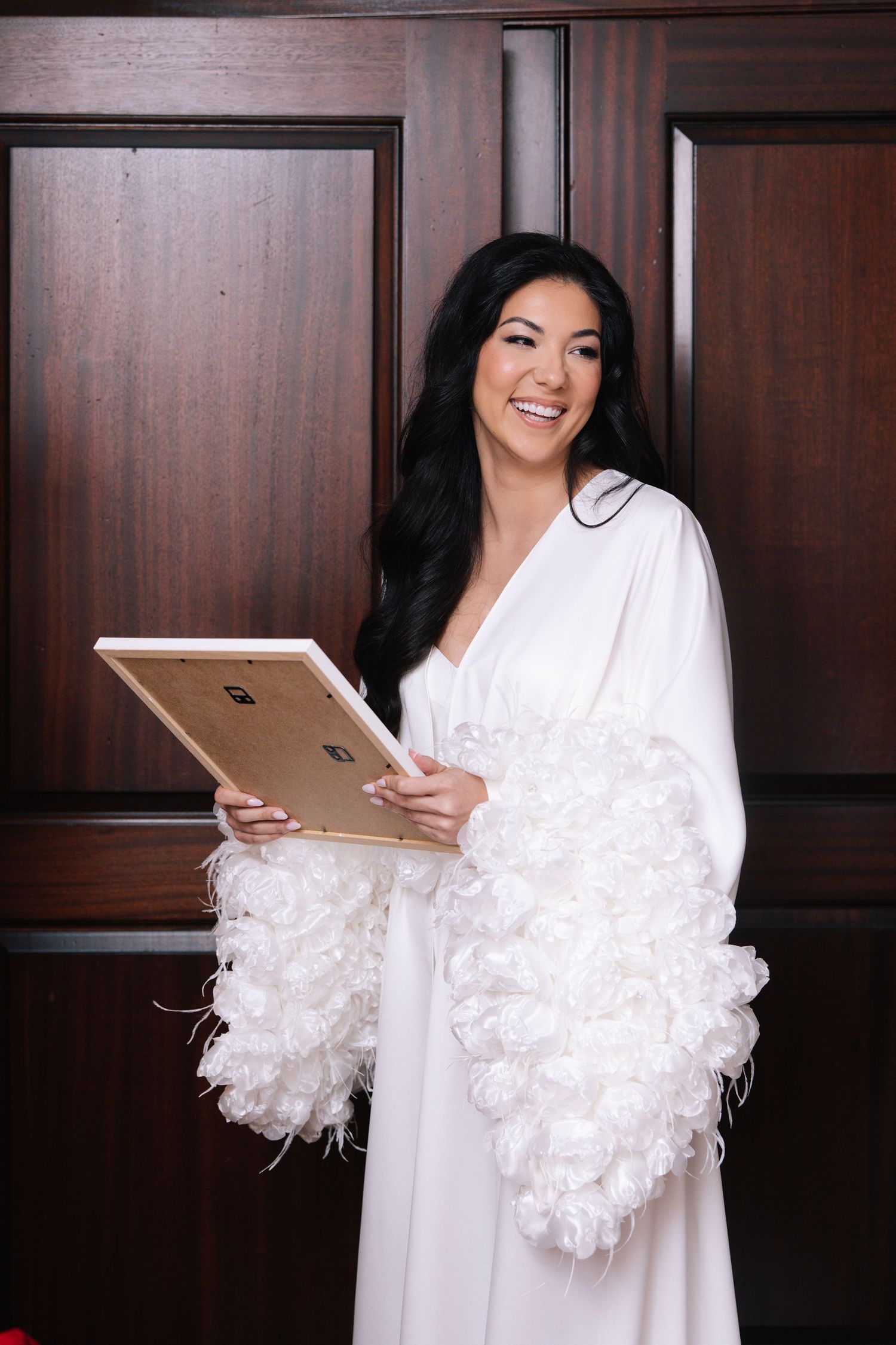 A person in a white robe with feathered sleeves holds a tablet device against a dark wood paneled wall.