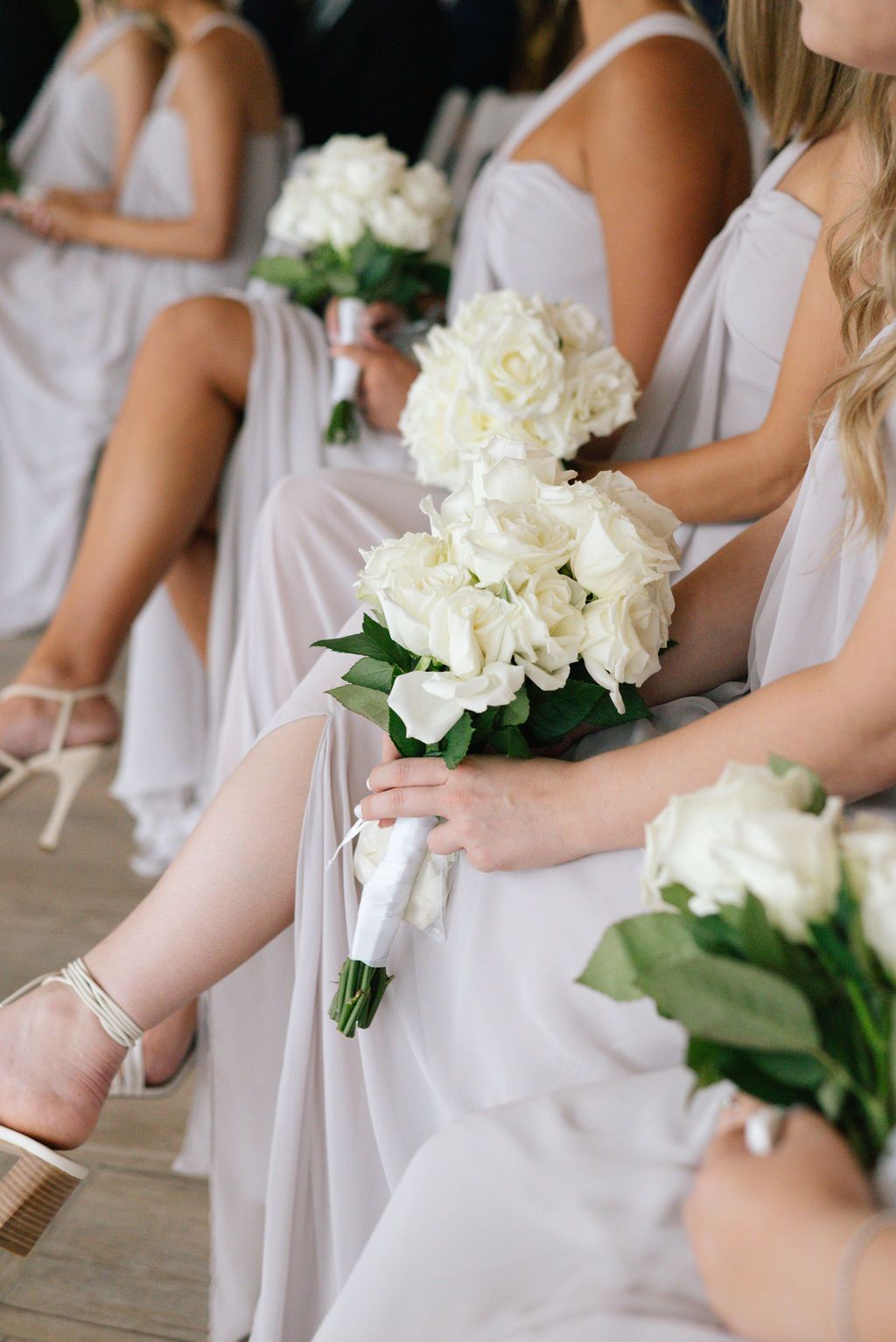 Row of bridesmaids in light dresses holding classic white rose bouquets.