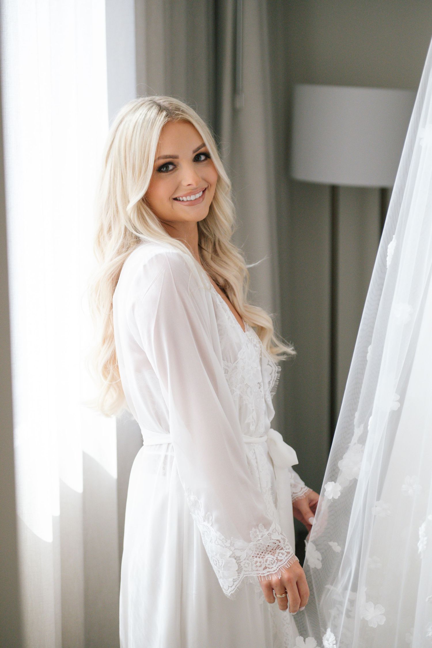 A bride in a white robe stands by a window during wedding day preparations.