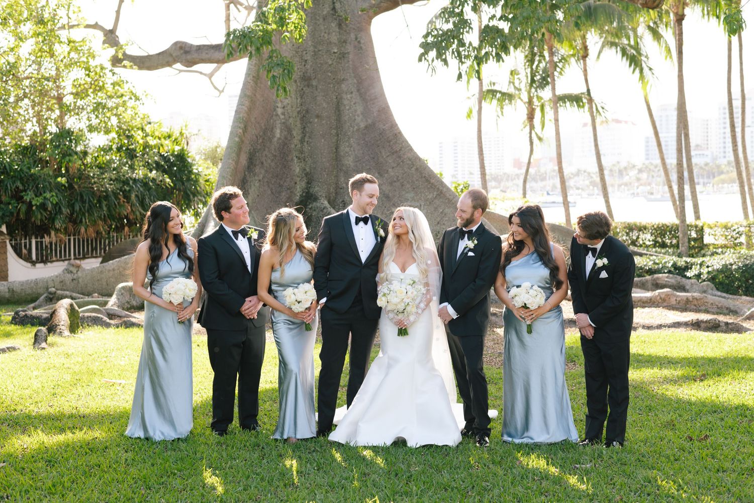 A wedding party poses together under large trees in a scenic outdoor garden setting.