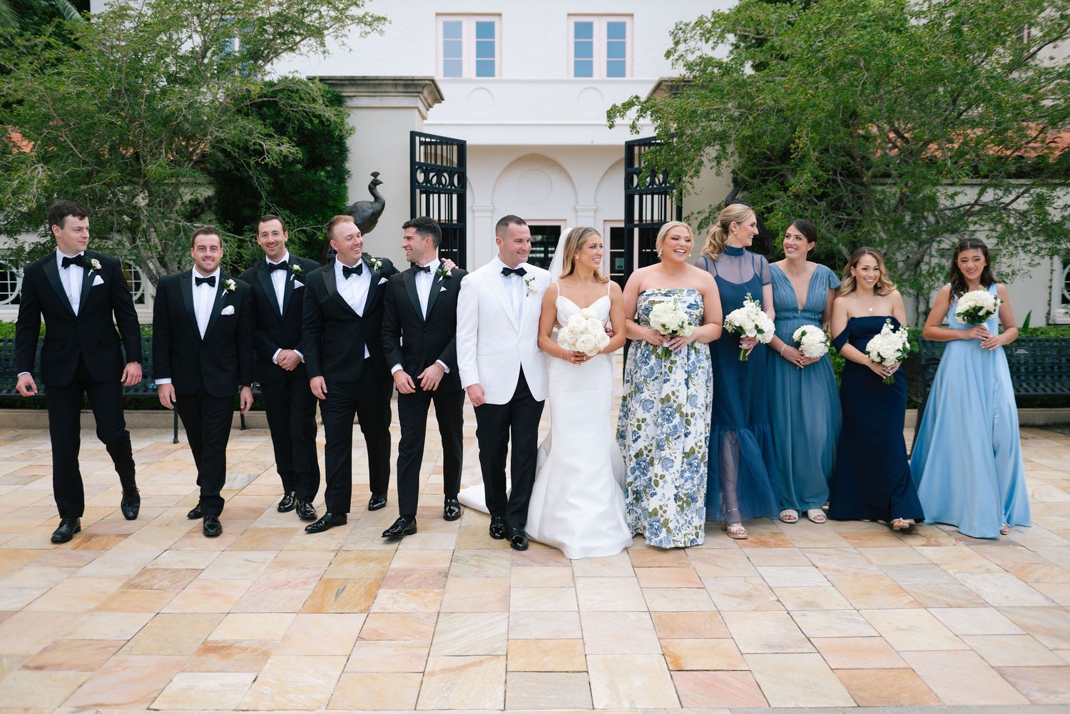 Wedding party in formal attire poses together on outdoor stone patio for group photos.