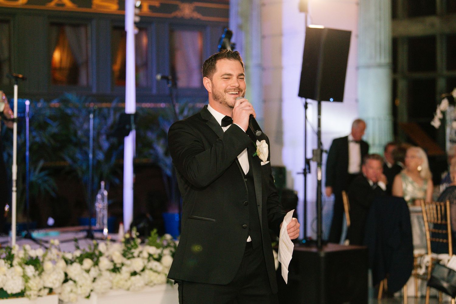 Dapper groom in black tuxedo delivers heartfelt speech at wedding reception.