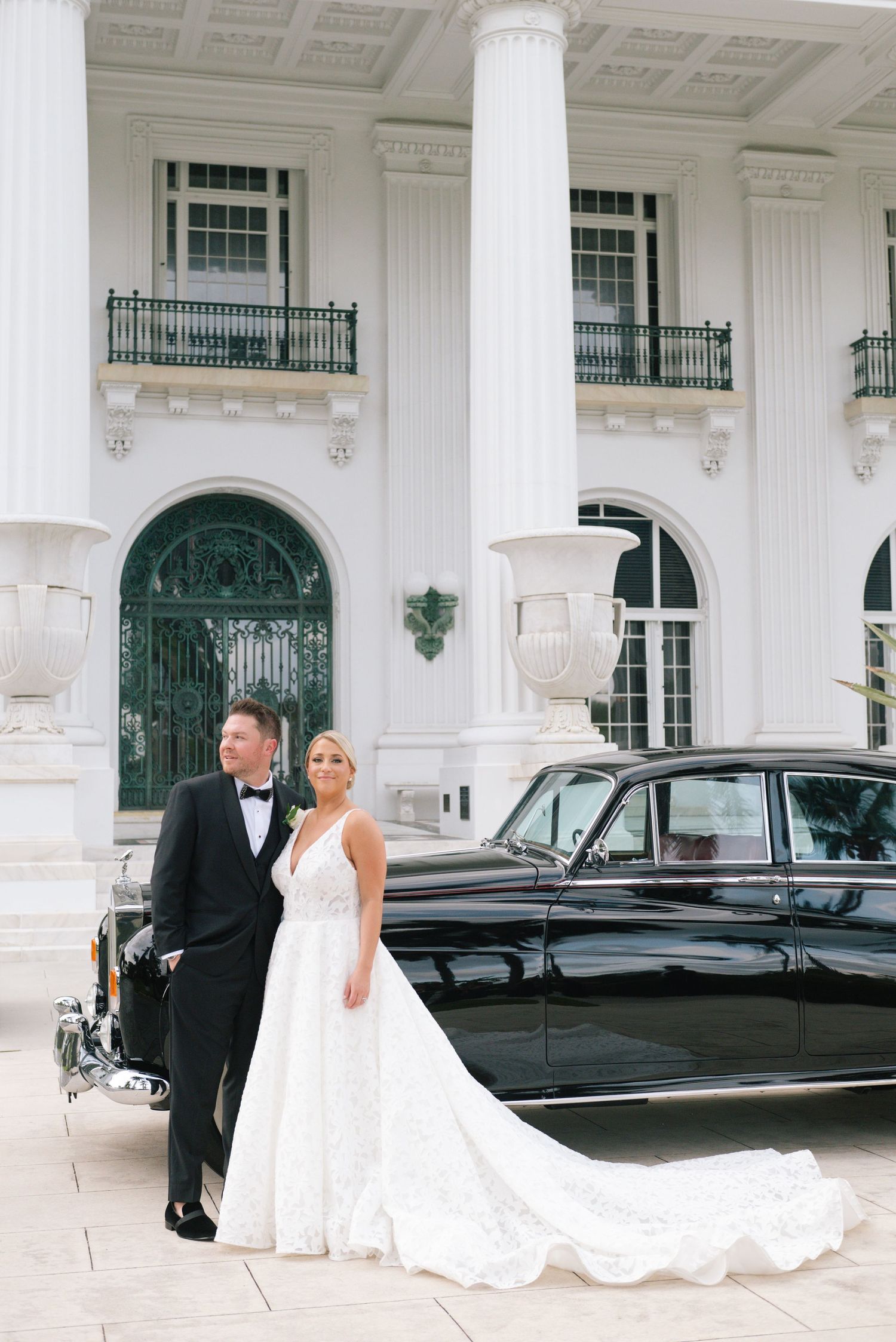 Bride and groom posing beside vintage black car in front of elegant white mansion.