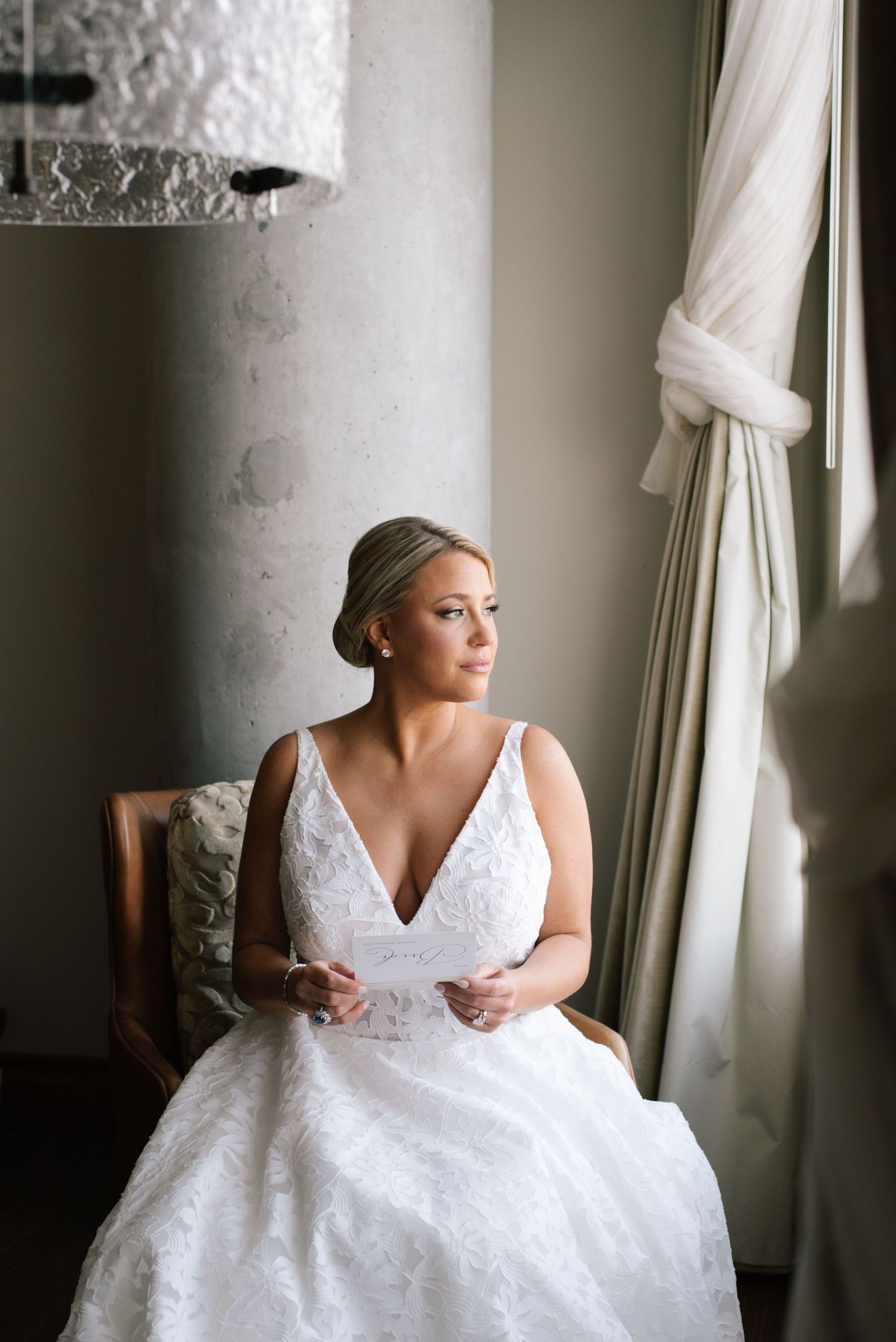 Elegant bride in white lace V-neck wedding gown seated by window in soft natural light.