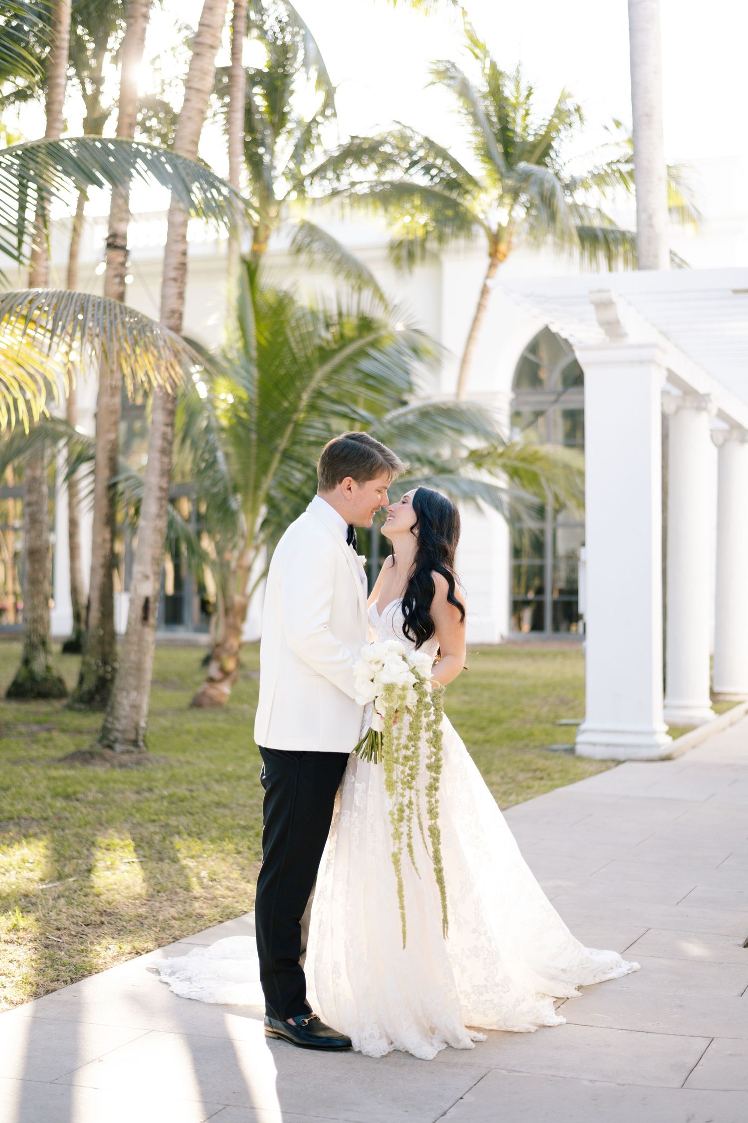 Romantic wedding photos captured under white columns with tropical palm trees in background.