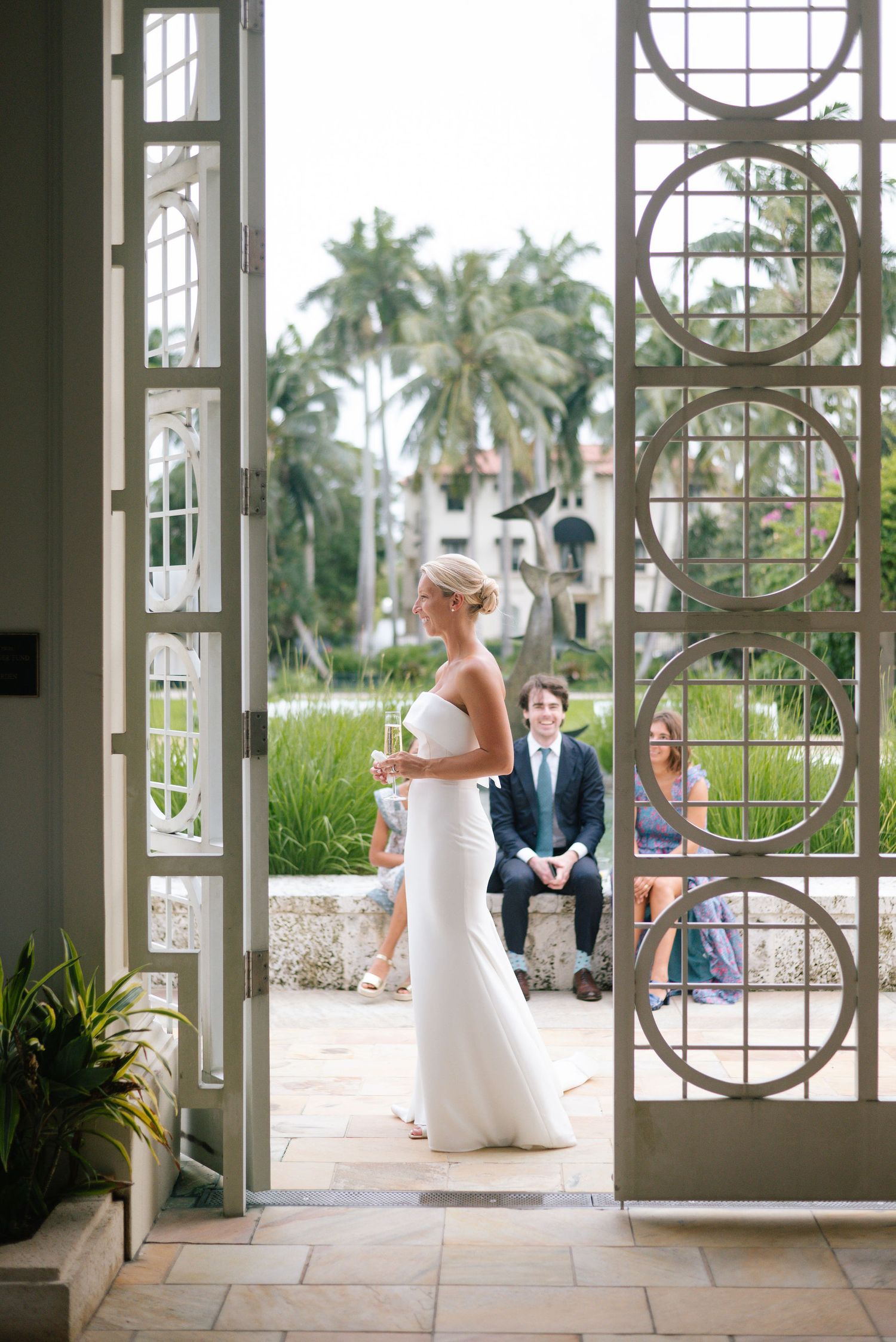 Bride in white gown standing by ornate glass doors with palm trees visible outside.