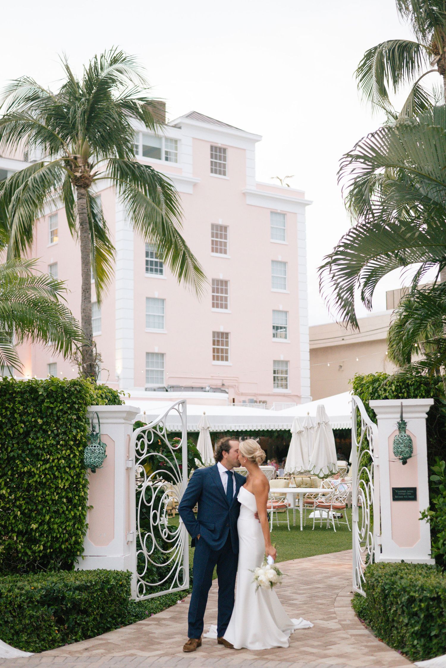 Elegant wedding couple poses along a tropical garden path with decorative white gates.