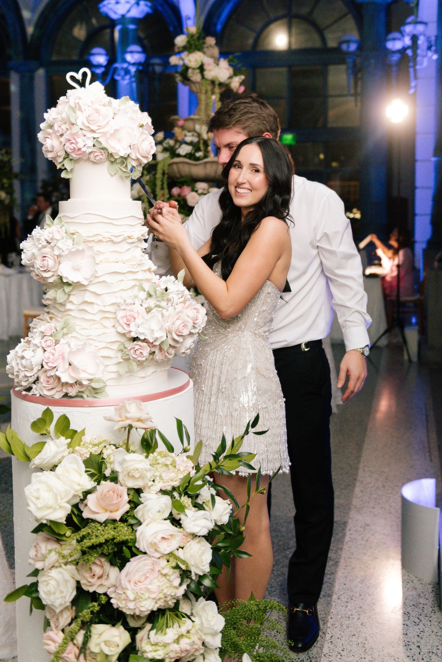 Elegant white wedding cake decorated with fresh flowers and greenery at a reception.