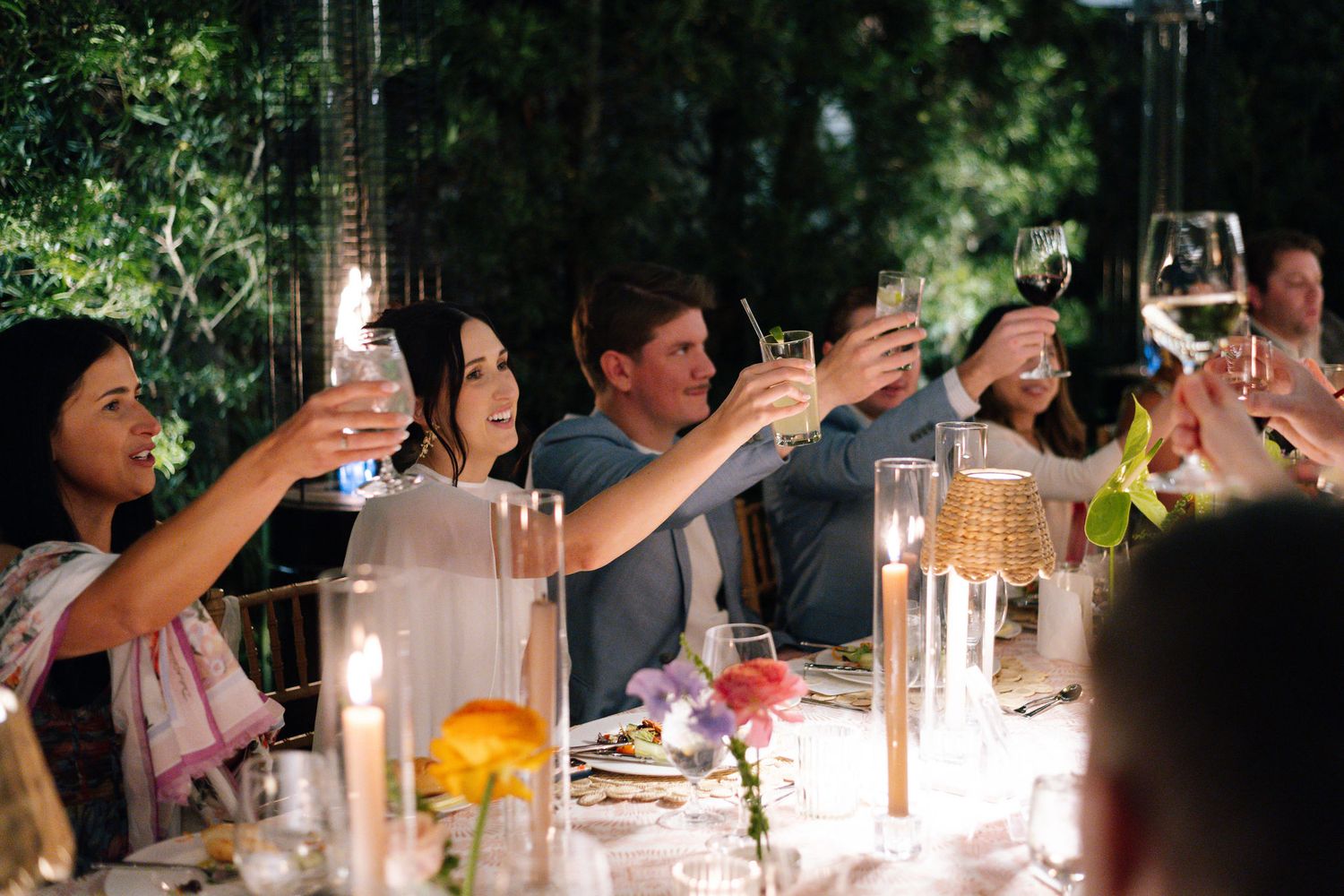 Wedding guests raise their glasses for a toast at an elegant candlelit dinner table.