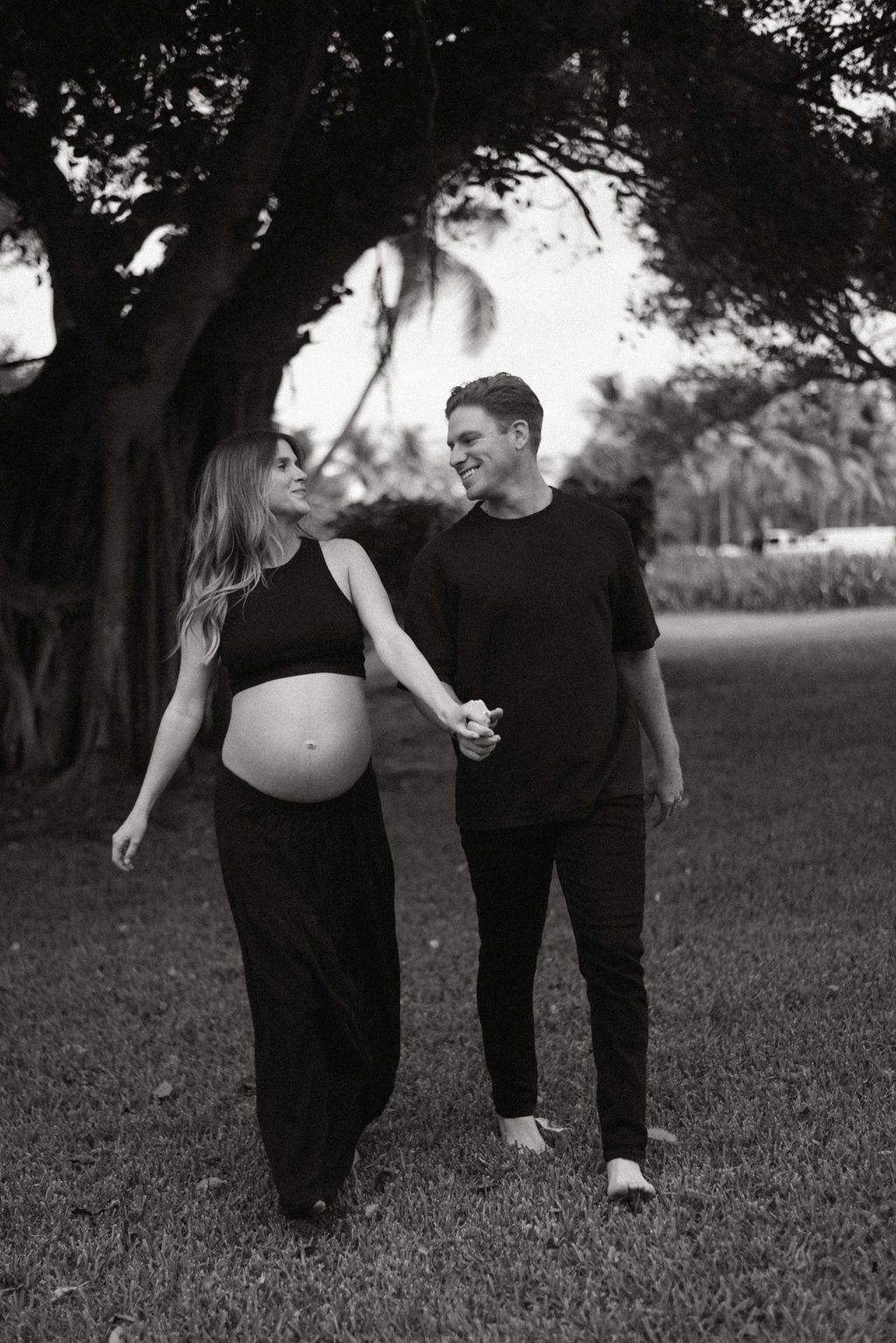 Black and white maternity photo of couple walking together through a grassy field.