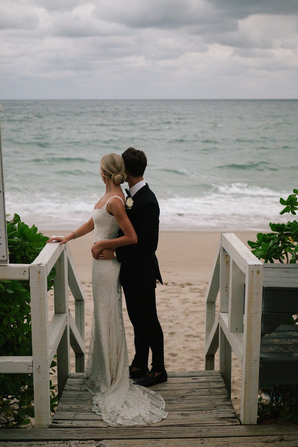 Wedding couple shares romantic moment on wooden boardwalk overlooking ocean.