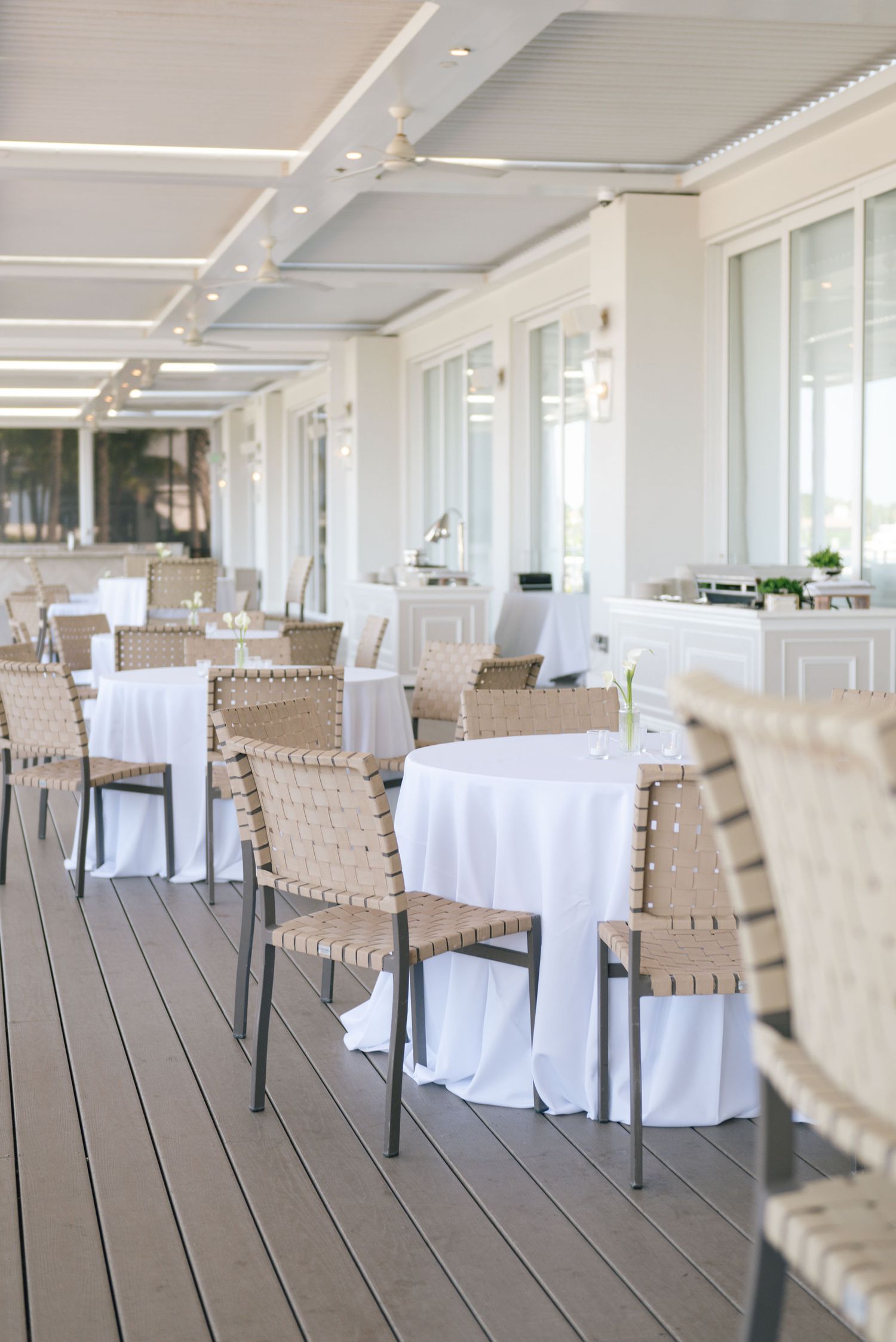 Modern coastal restaurant interior with white tables and woven chairs on wooden deck.