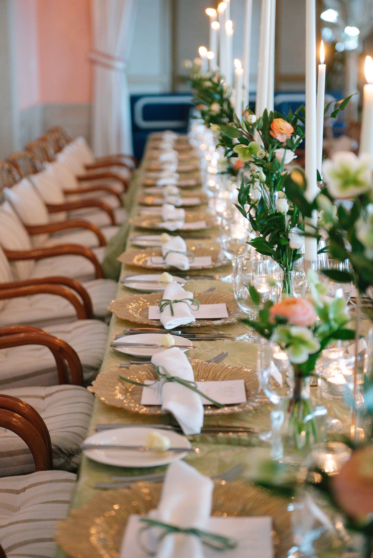 Long banquet table with gold chargers, white napkins and floral arrangements featuring tall white candles.
