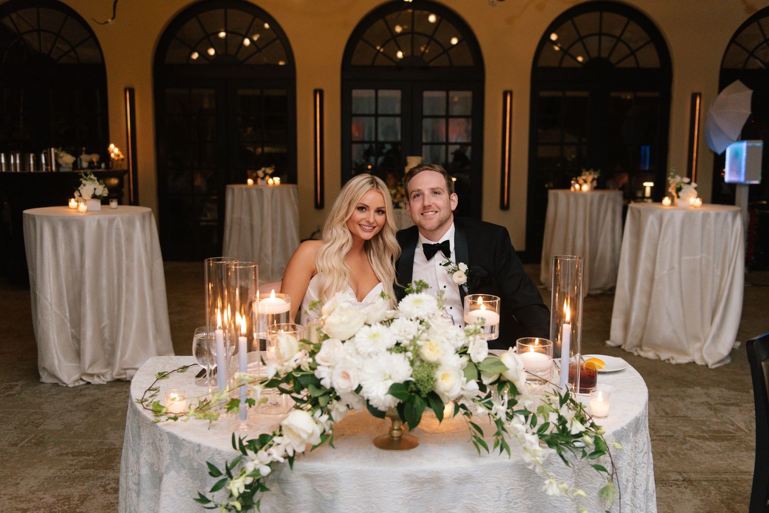 An elegant wedding reception setup with white floral centerpieces and glowing candles on dining tables.