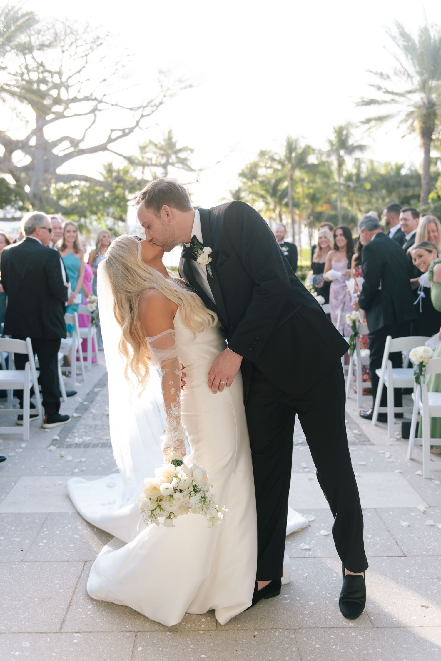 Wedding couple shares romantic moments during their outdoor ceremony with palm trees and white chairs in background.