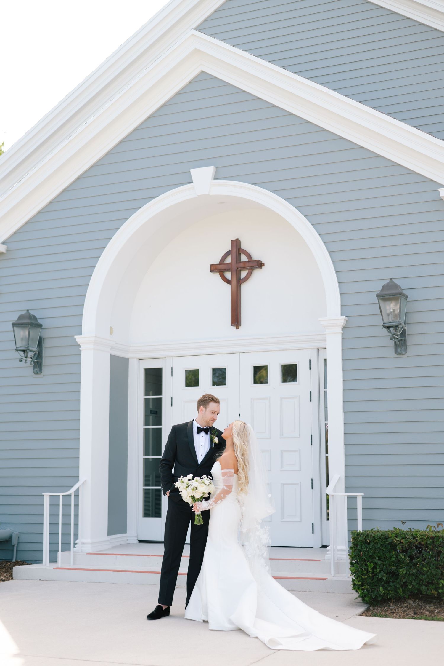 A romantic wedding couple poses in front of a white chapel with elegant arched doorways and grey siding.