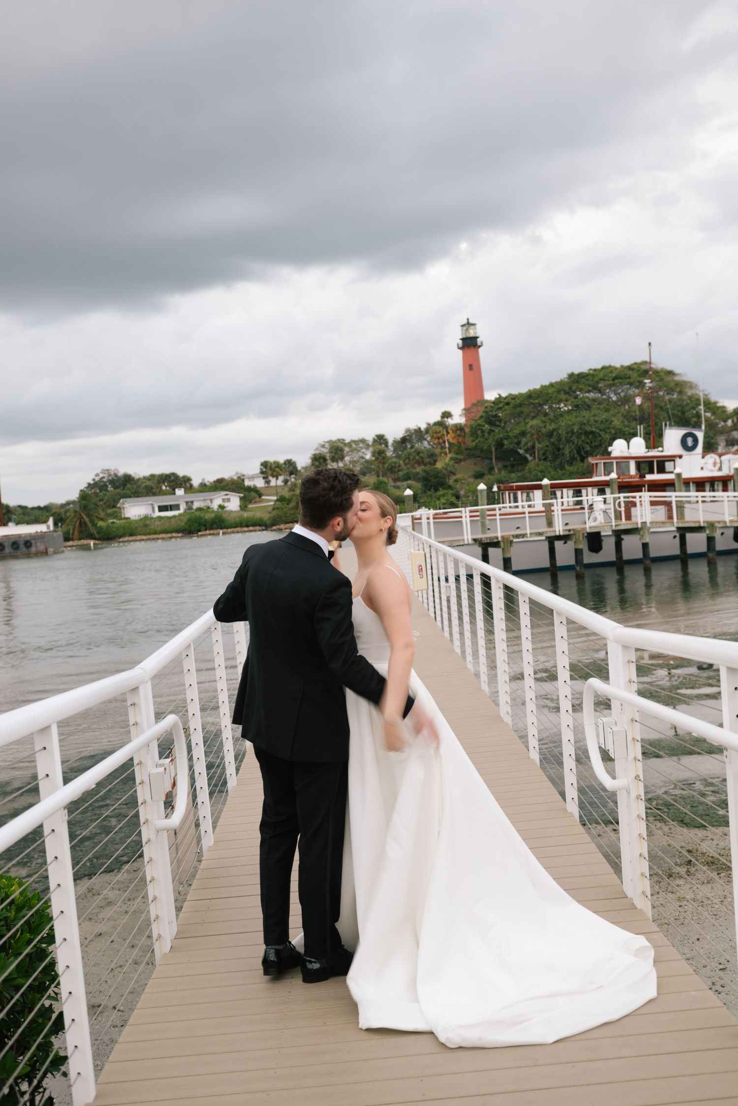 Wedding couple on boardwalk with lighthouse and cloudy sky in background.