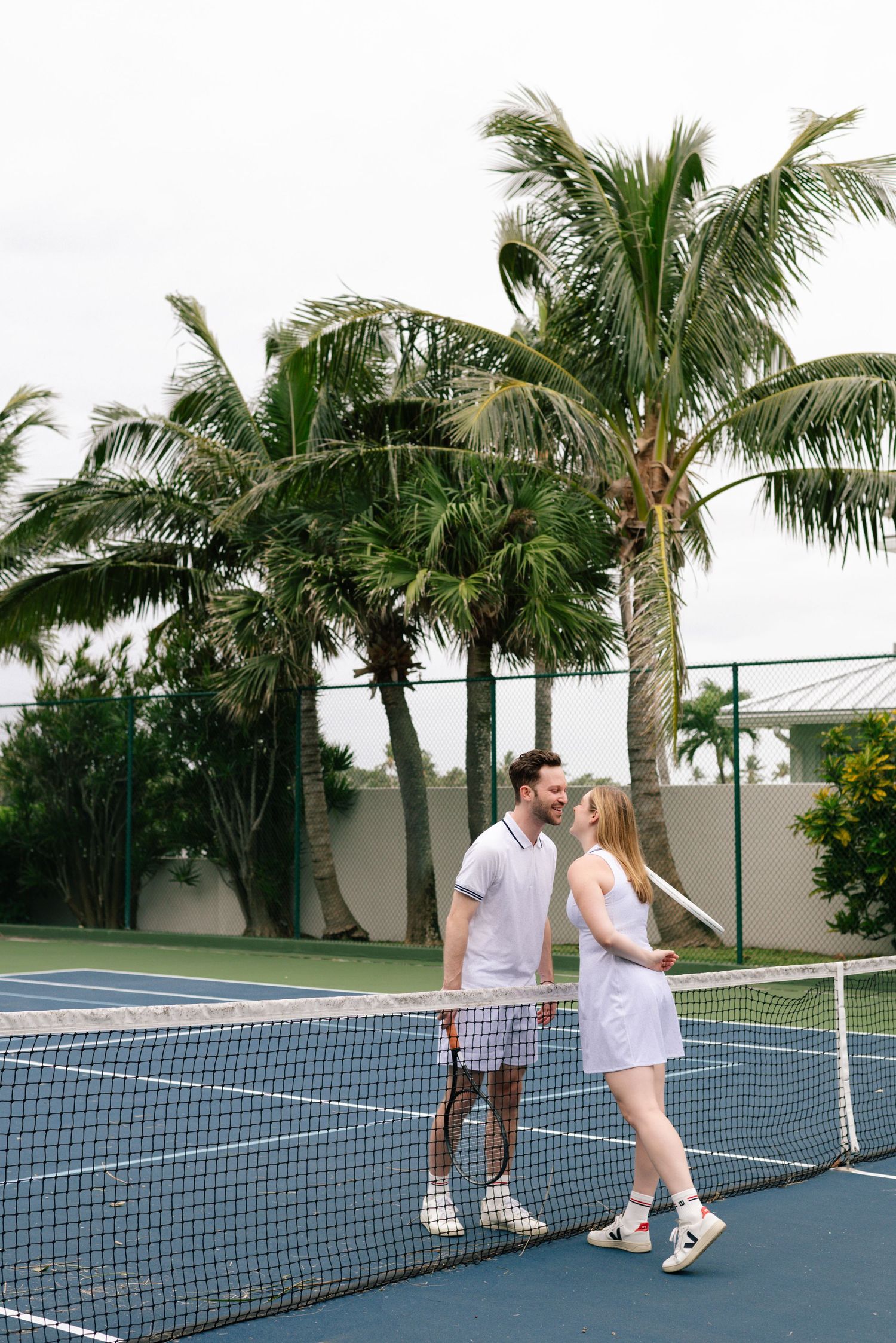 A couple in white tennis outfits stands by the net on a blue court surrounded by palm trees.