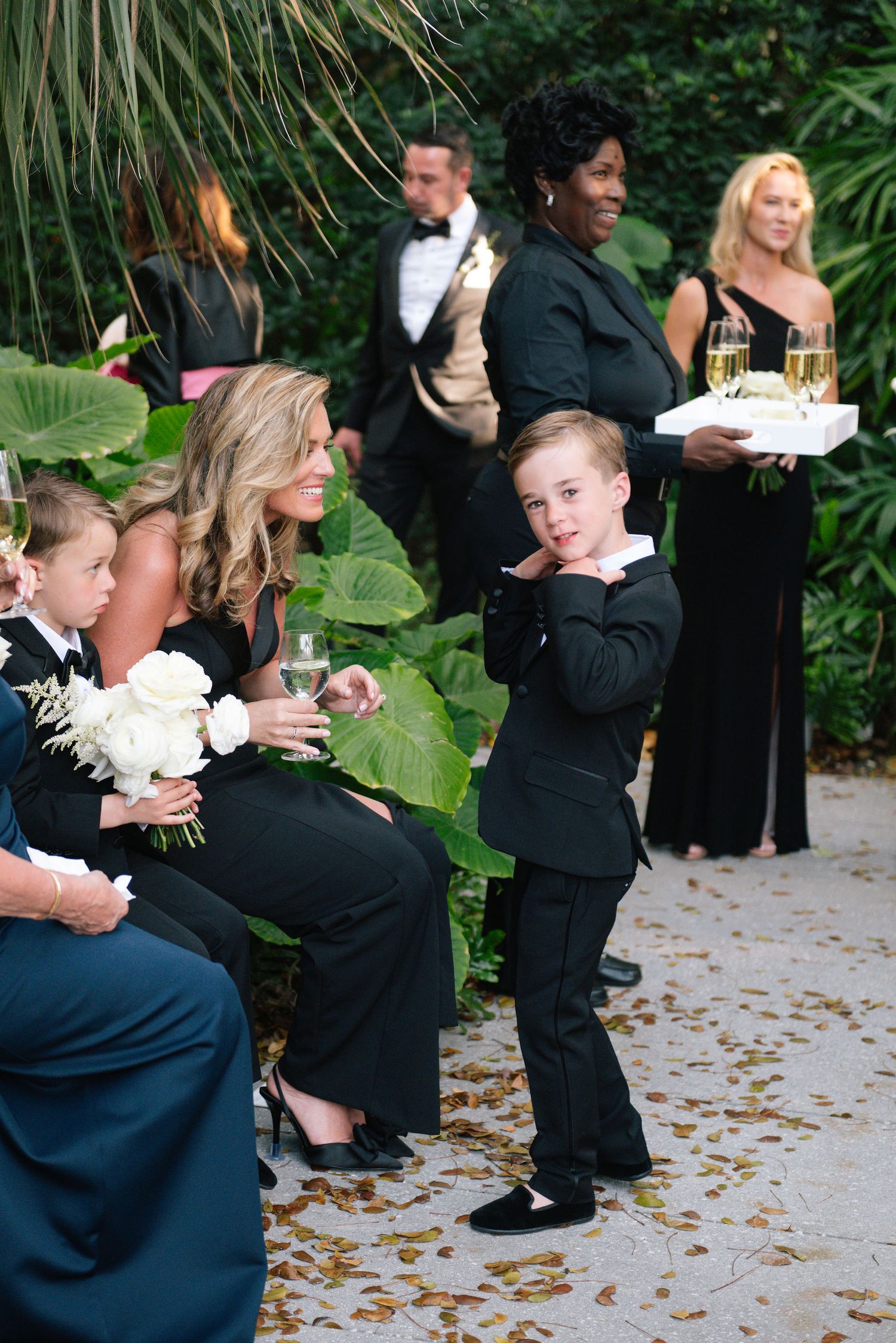 Wedding guests in formal black attire dance and celebrate on a stone patio surrounded by greenery.
