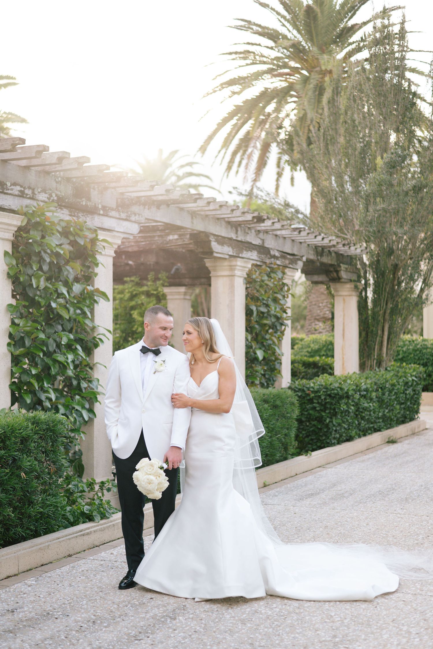 Newlyweds walking along a palm-lined pathway with white columns at a luxury venue.