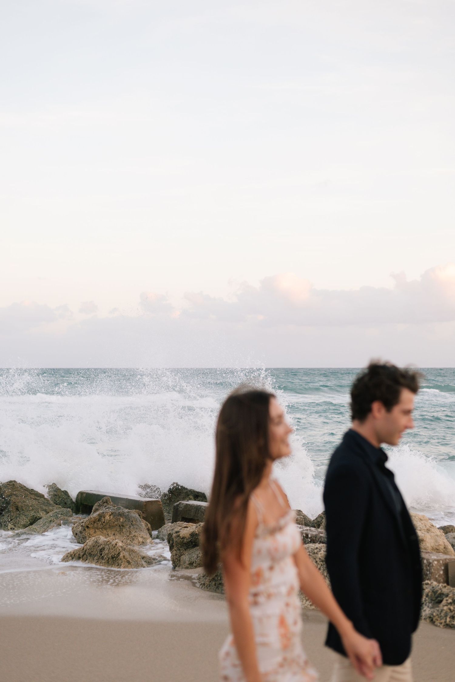 Two people embrace by the ocean as waves crash against rocky outcrops during sunset.