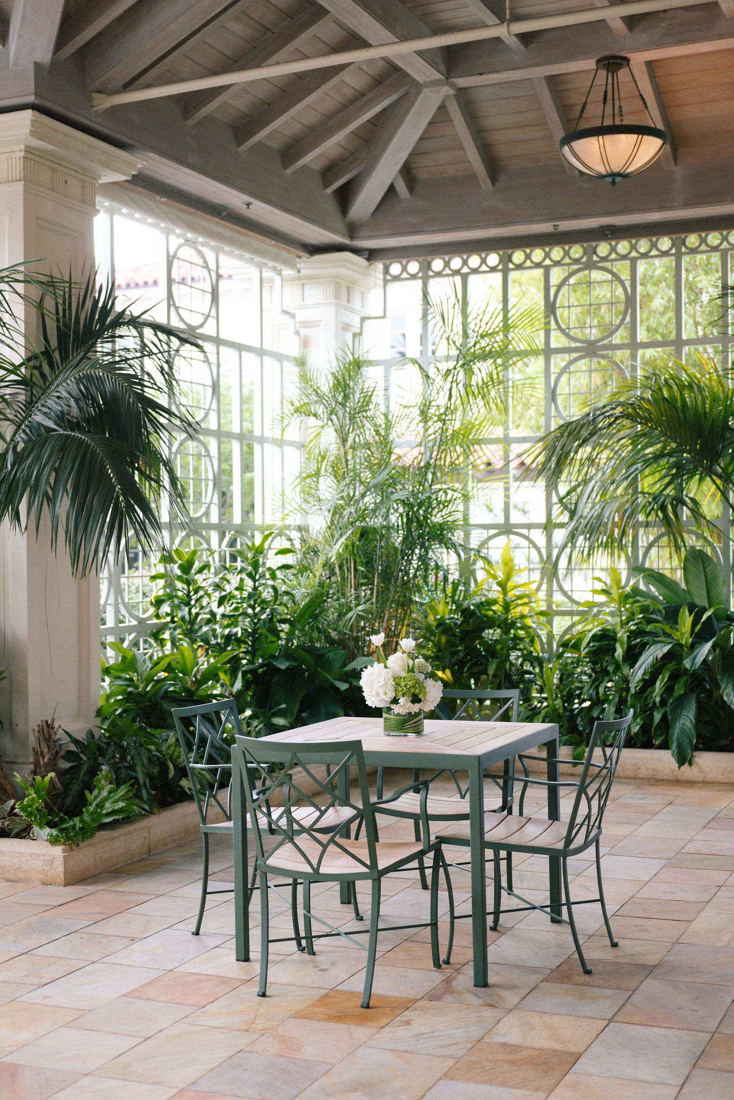 Sunlit conservatory dining area with green metal chairs and tropical plants.