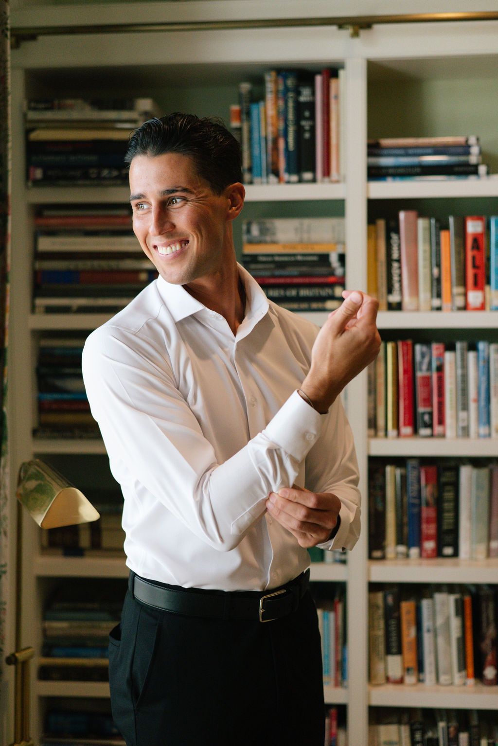 Person in white dress shirt adjusting cufflinks in front of bookshelf.