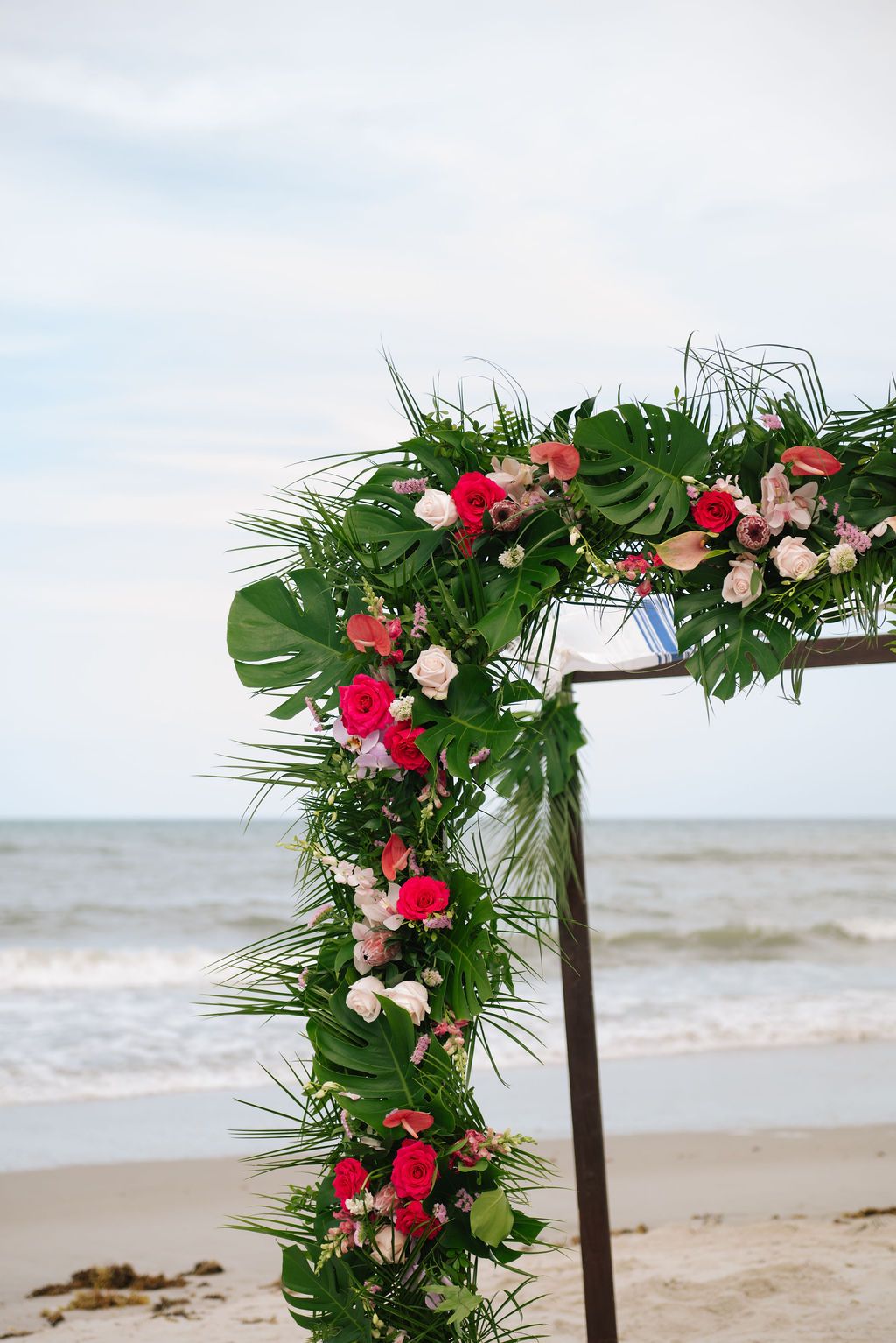 Wooden wedding arch decorated with pink flowers and tropical greenery on a beach.