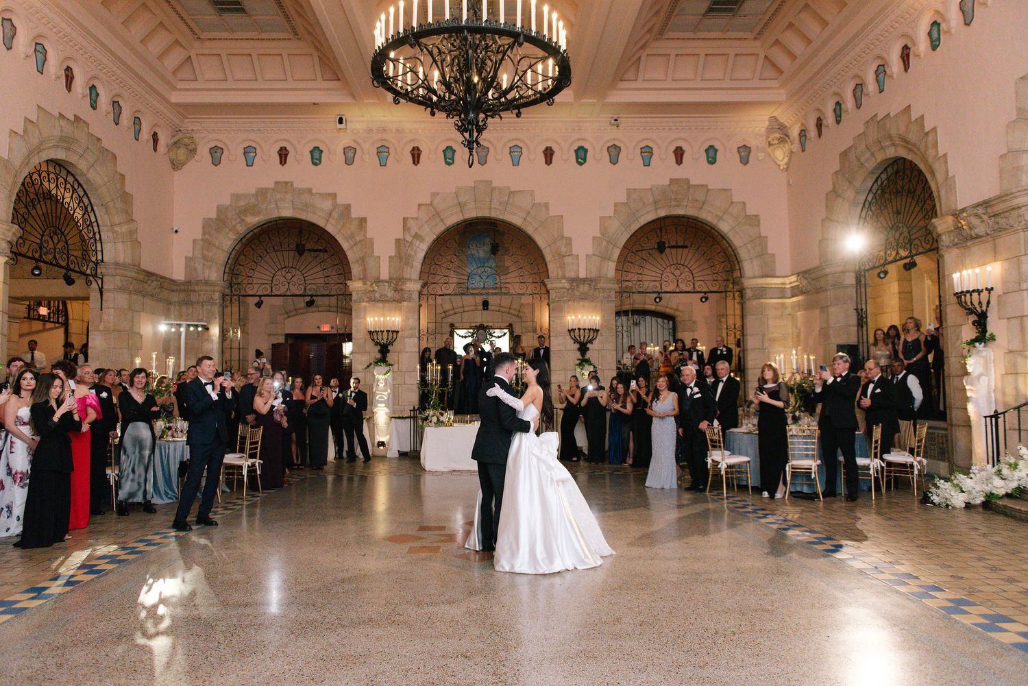 Grand ballroom with ornate chandelier and arched architecture filled with wedding guests.