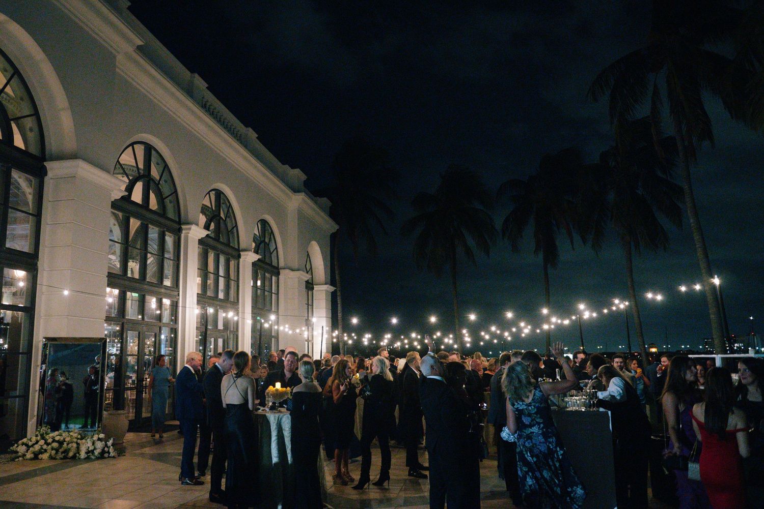 Guests mingle outside elegant white venue with palm trees and string lights at night.