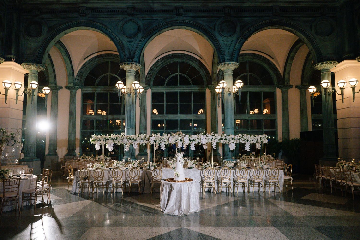 Elegant wedding reception hall featuring tall floral centerpieces, chandeliers, and gold chiavari chairs.