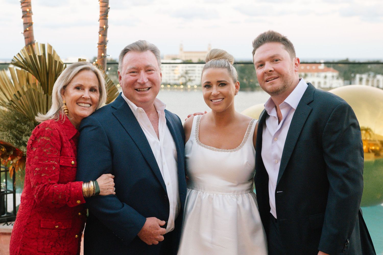 A celebratory group photo at an outdoor venue with palm trees and water in the background.