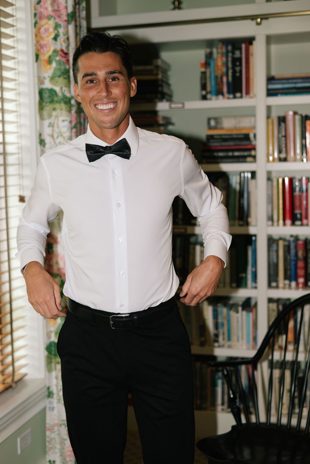 Person in white shirt and bow tie smiling in front of bookshelf.