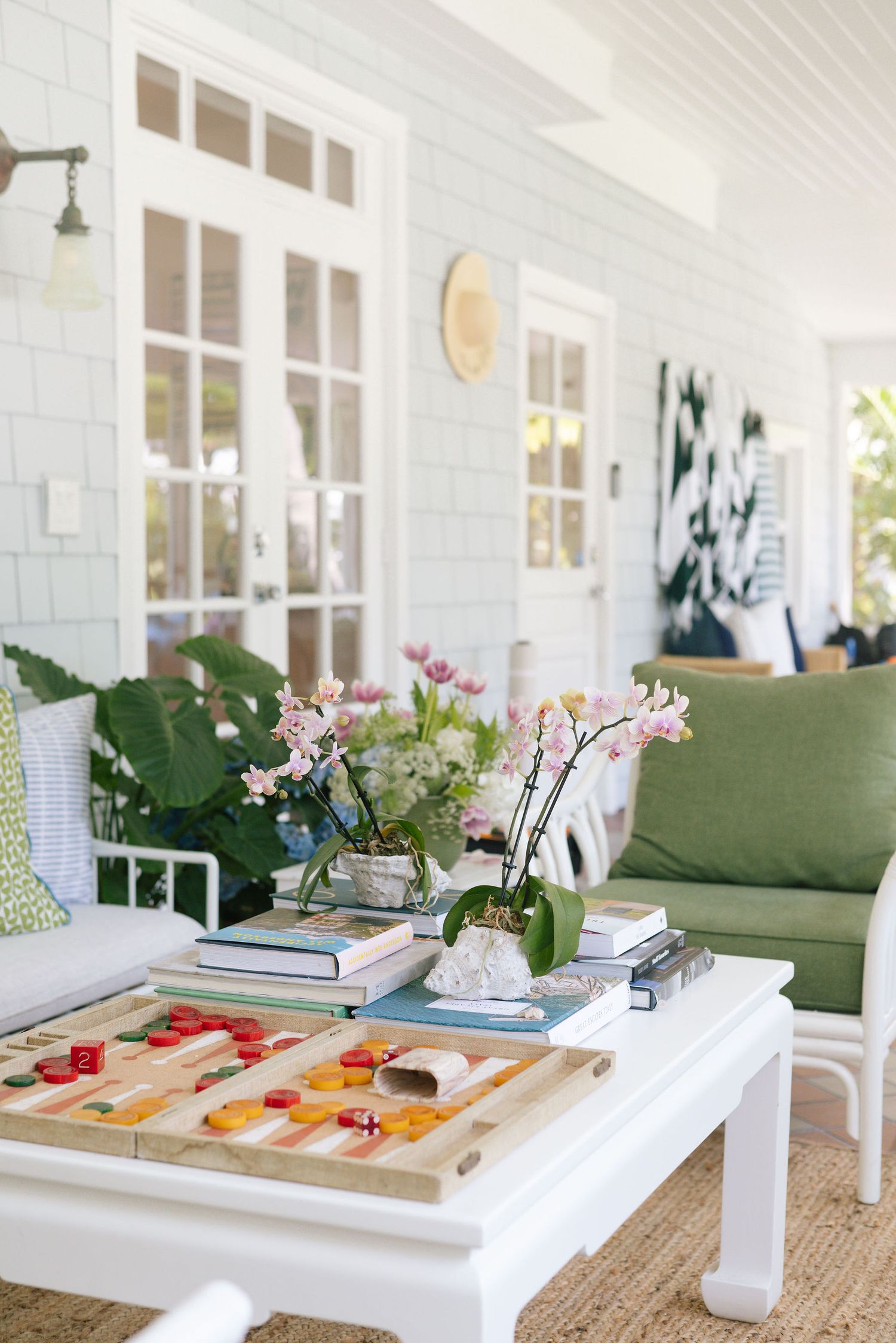 Bright sunlit porch interior with green sofa, orchids, and board games on white coffee table.