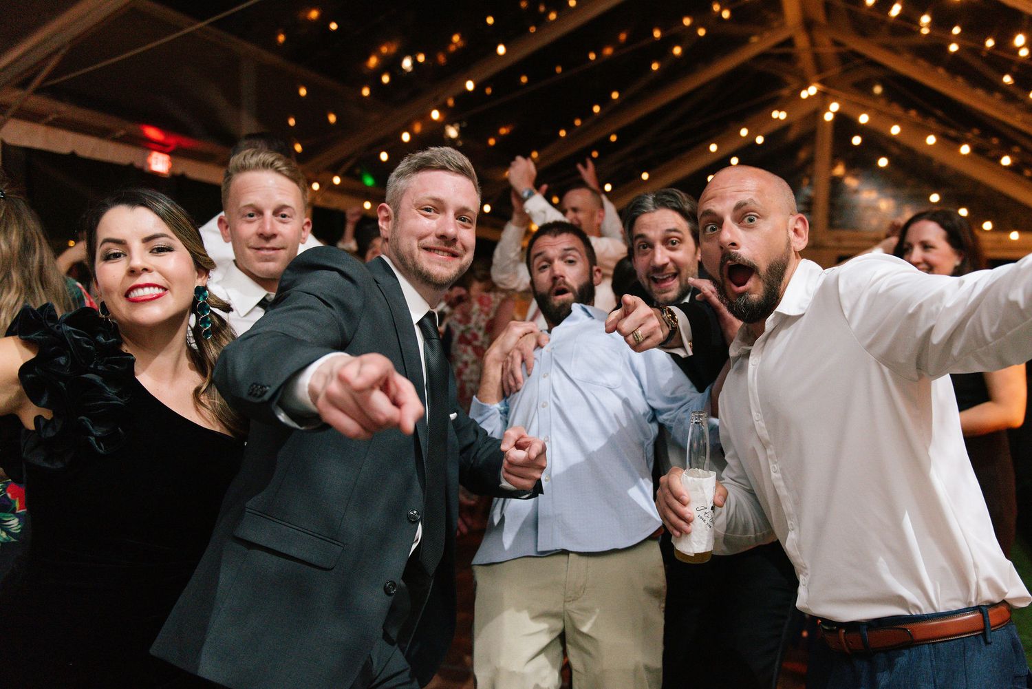 Wedding reception dance floor filled with guests under warm string lights and tent covering.