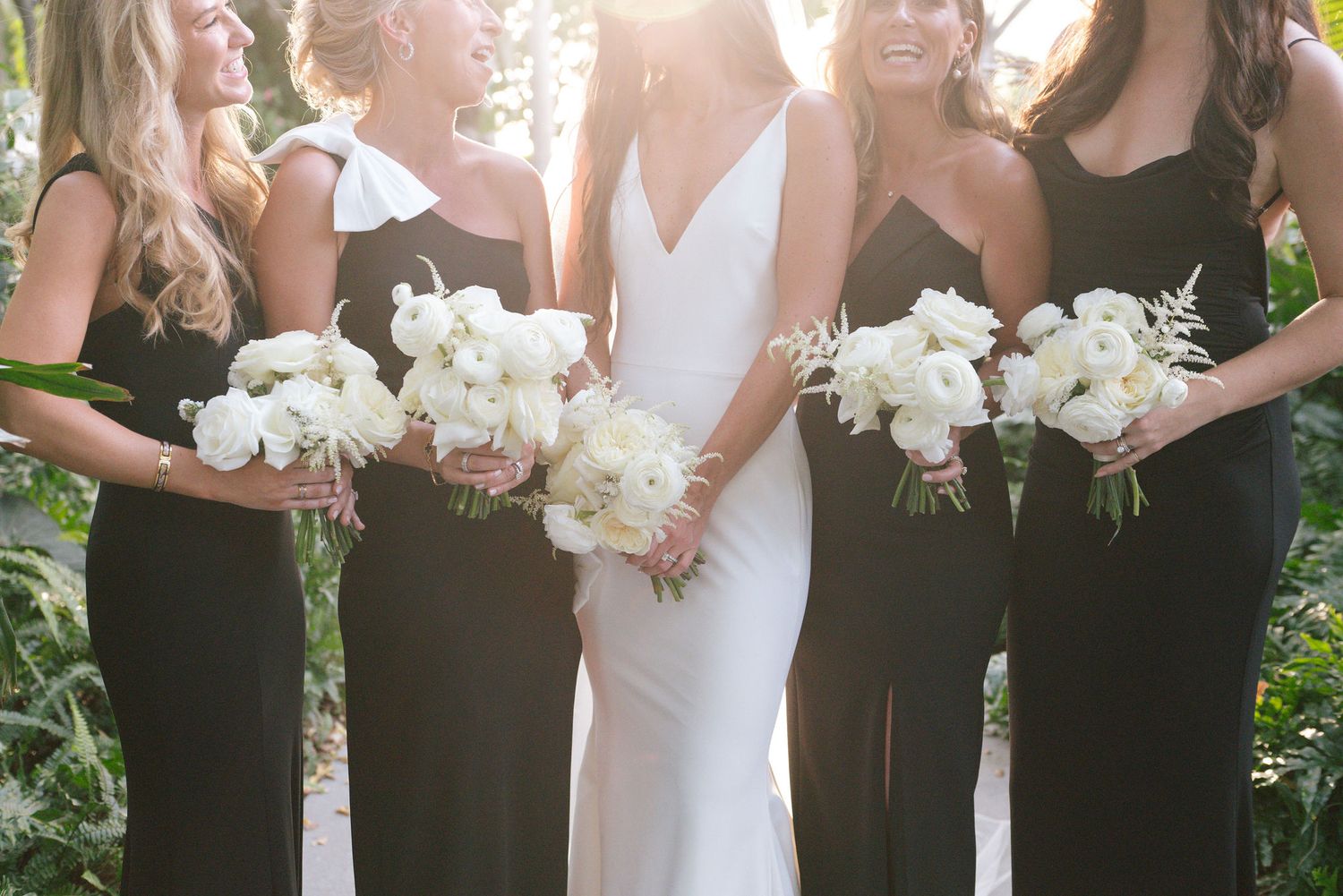 Wedding party wearing black bridesmaid dresses holding white floral bouquets stand in line against outdoor backdrop.