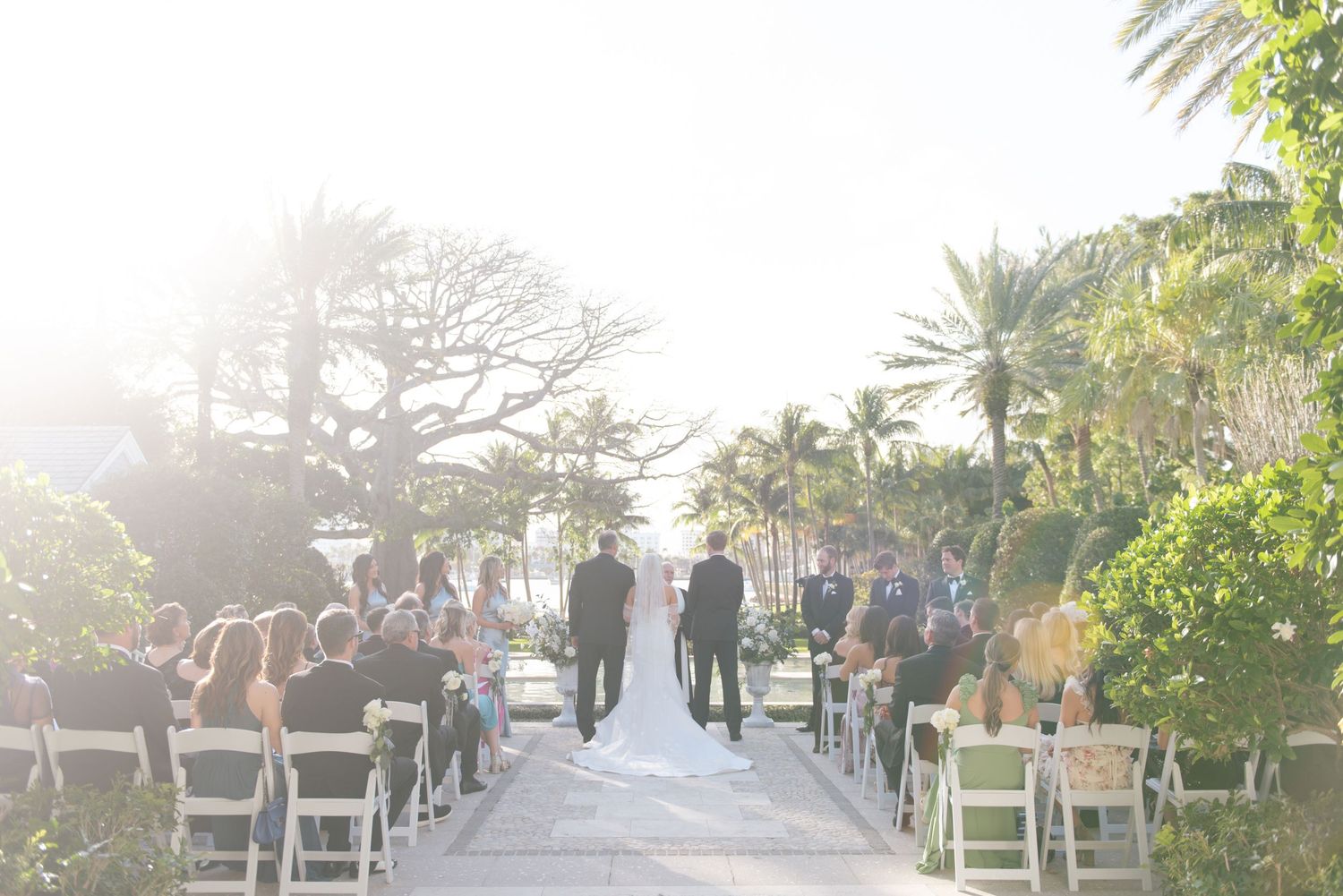 Outdoor wedding ceremony setup with white chairs arranged in rows under palm trees at sunset.