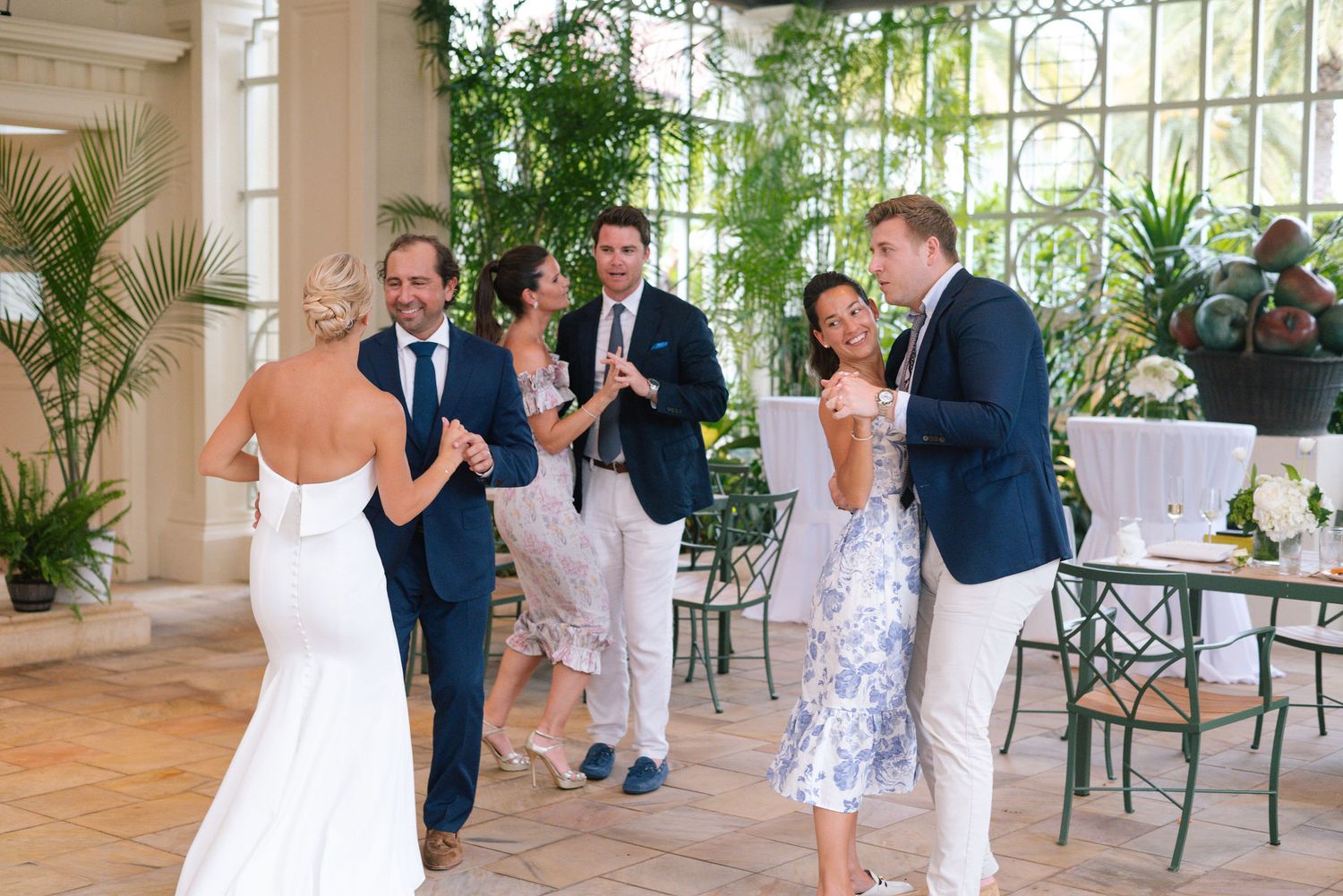 Wedding celebration group gathered in a bright conservatory with palm trees and natural light.