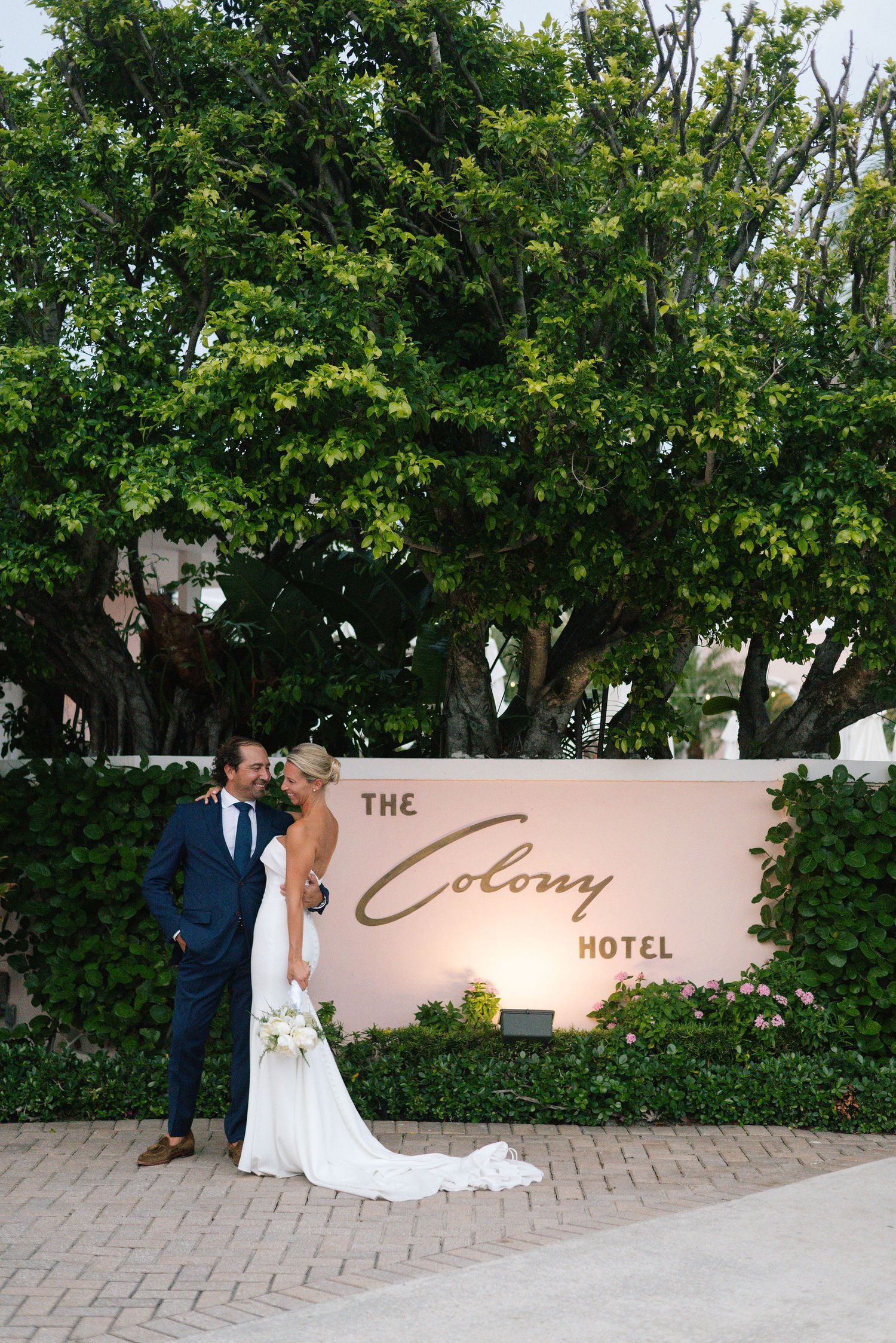 Wedding couple poses by The Colony Hotel sign surrounded by lush greenery.