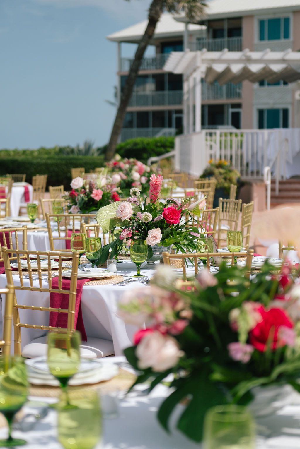 Outdoor wedding reception setup with gold chairs and pink floral centerpieces by the beach.