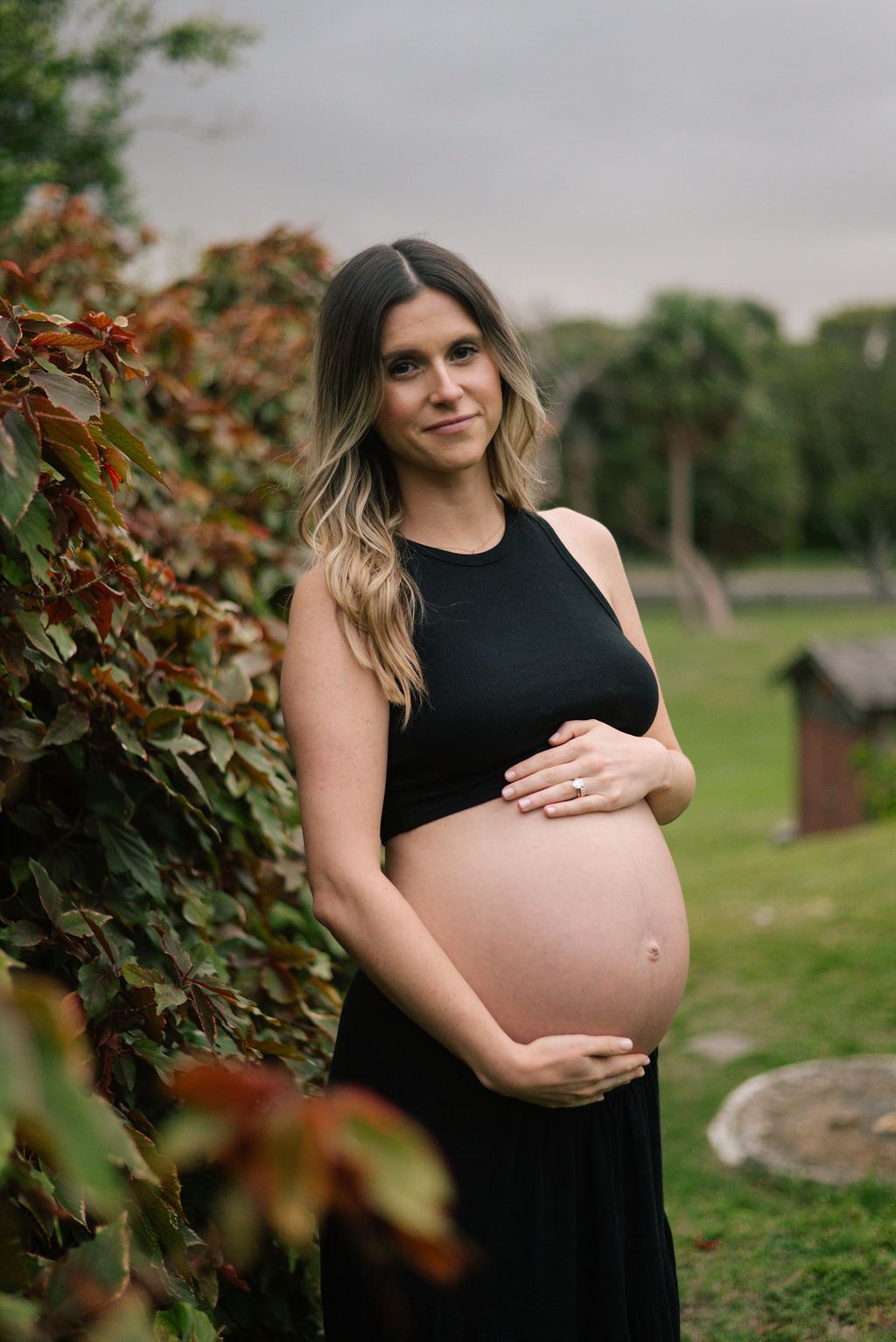 Maternity photoshoot in a garden setting featuring a black dress and natural outdoor backdrop.