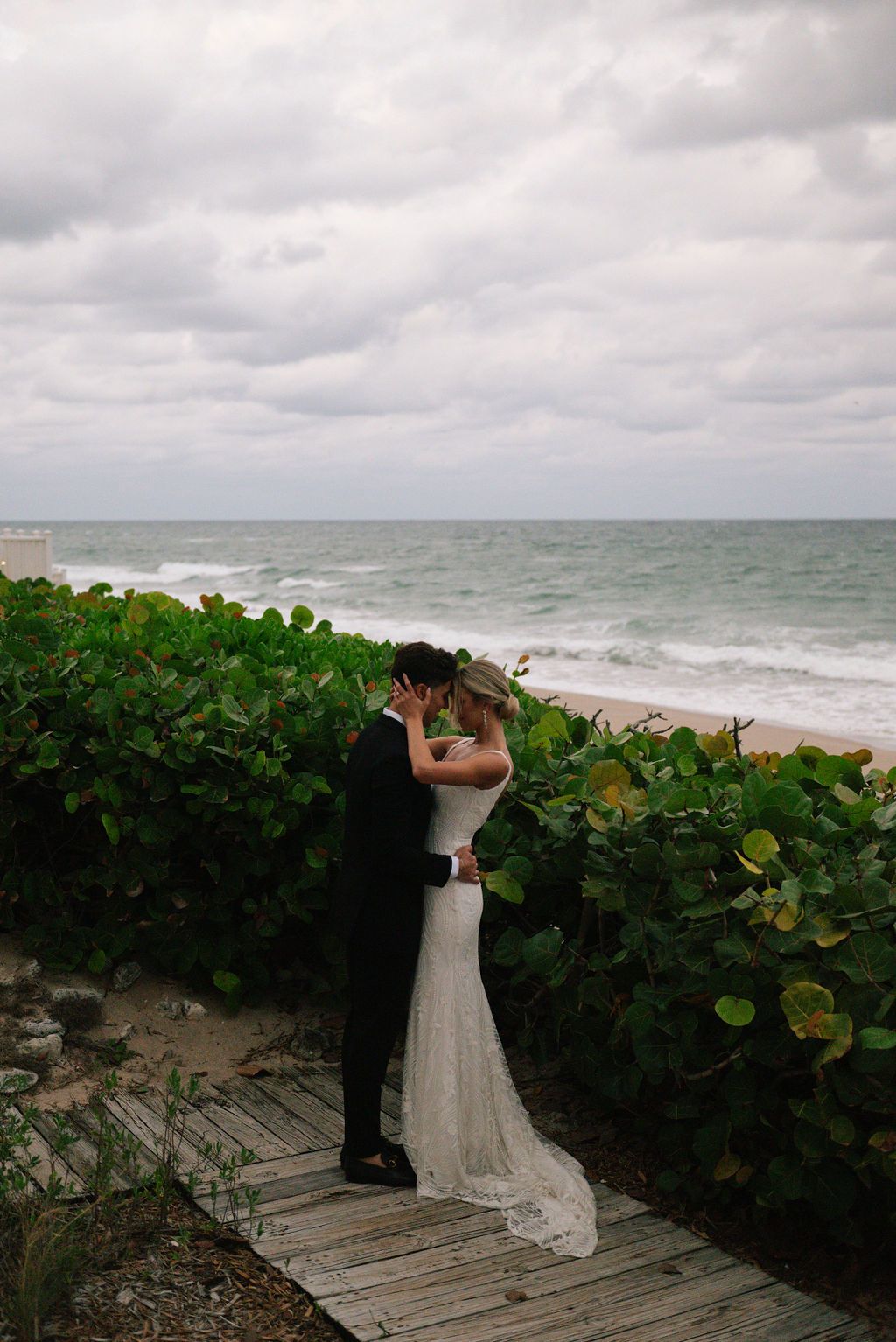 Wedding couple poses on wooden walkway with ocean and cloudy sky in background.
