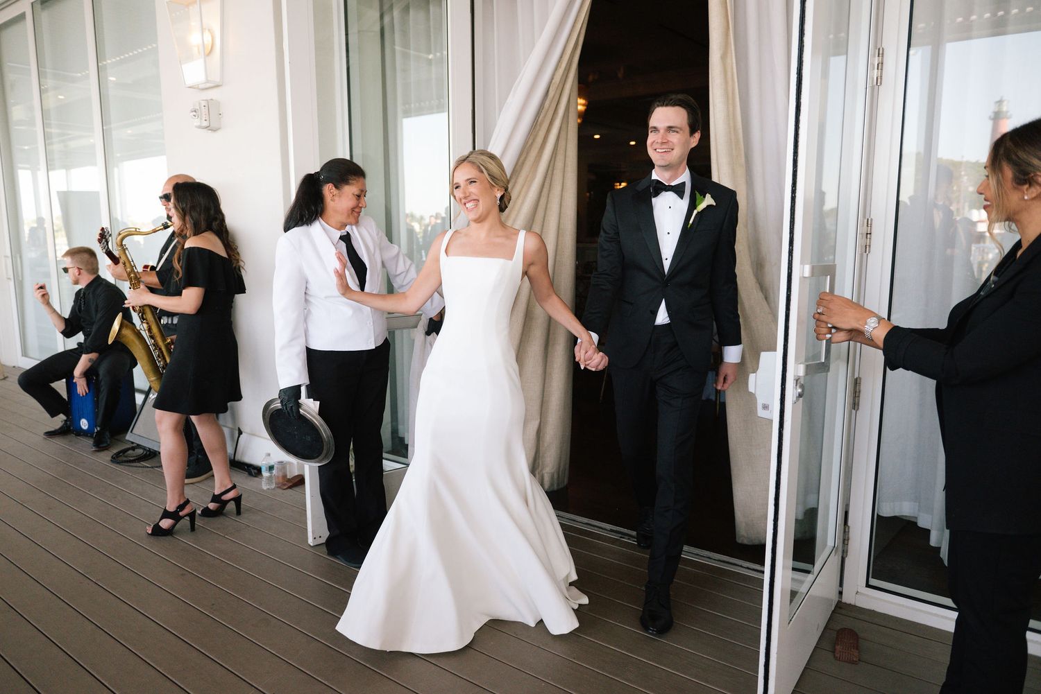 Newlyweds exit wedding venue onto outdoor deck surrounded by guests.