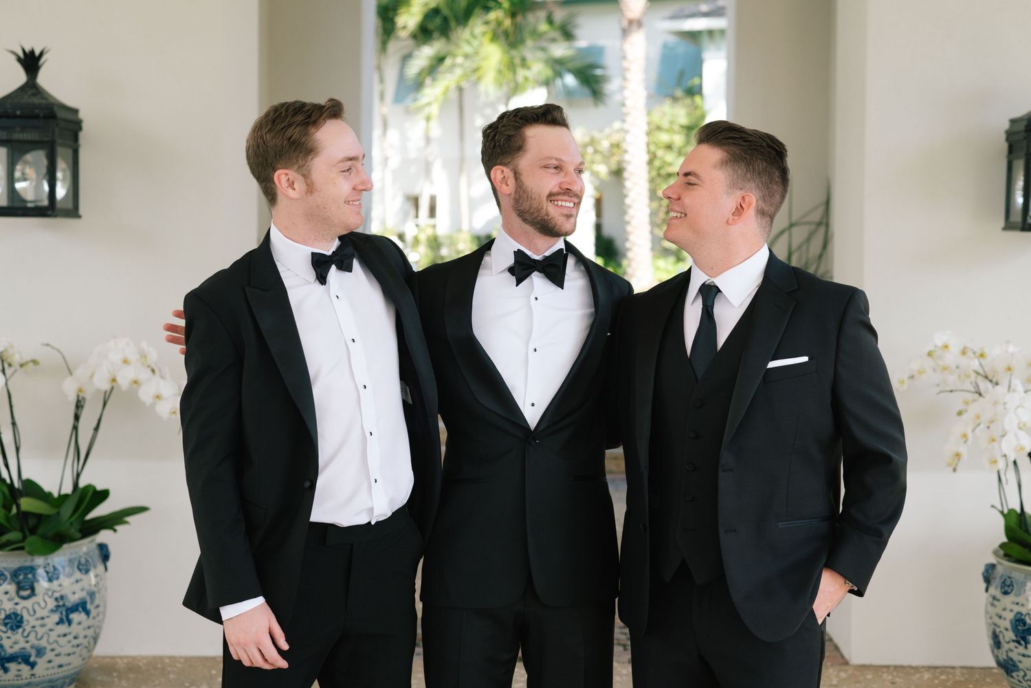 Three groomsmen in tuxedos sharing a laugh in front of white pillars.