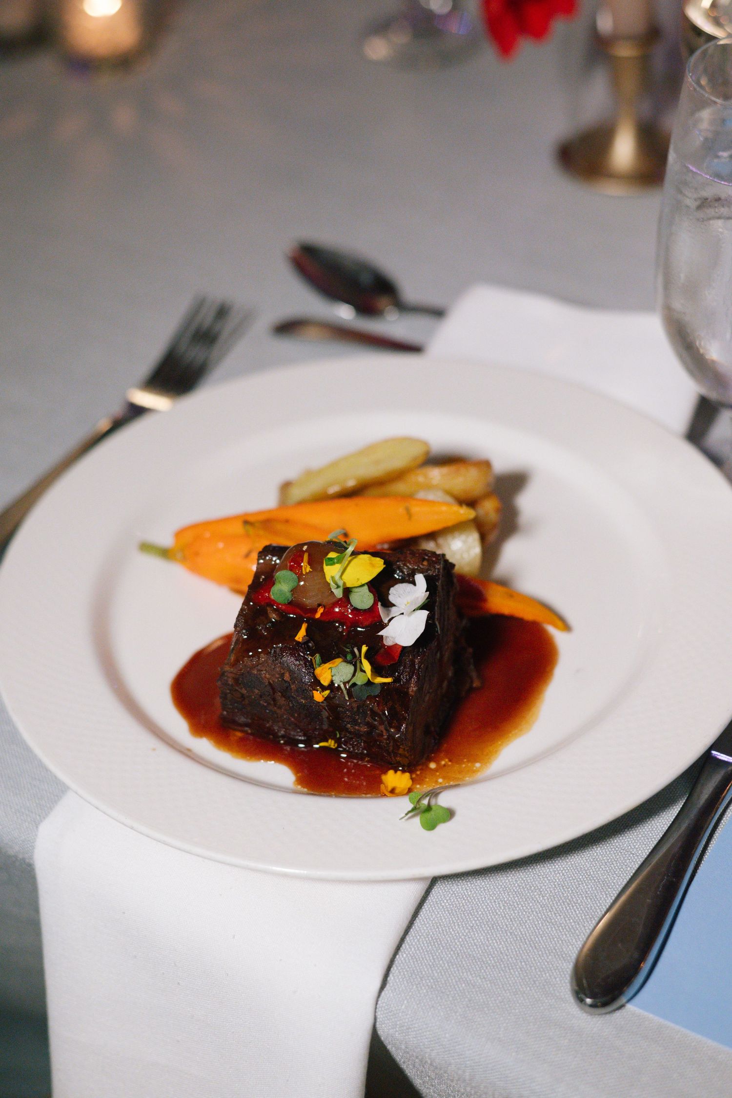 Plated dinner service featuring braised short rib with colorful garnishes on white china.