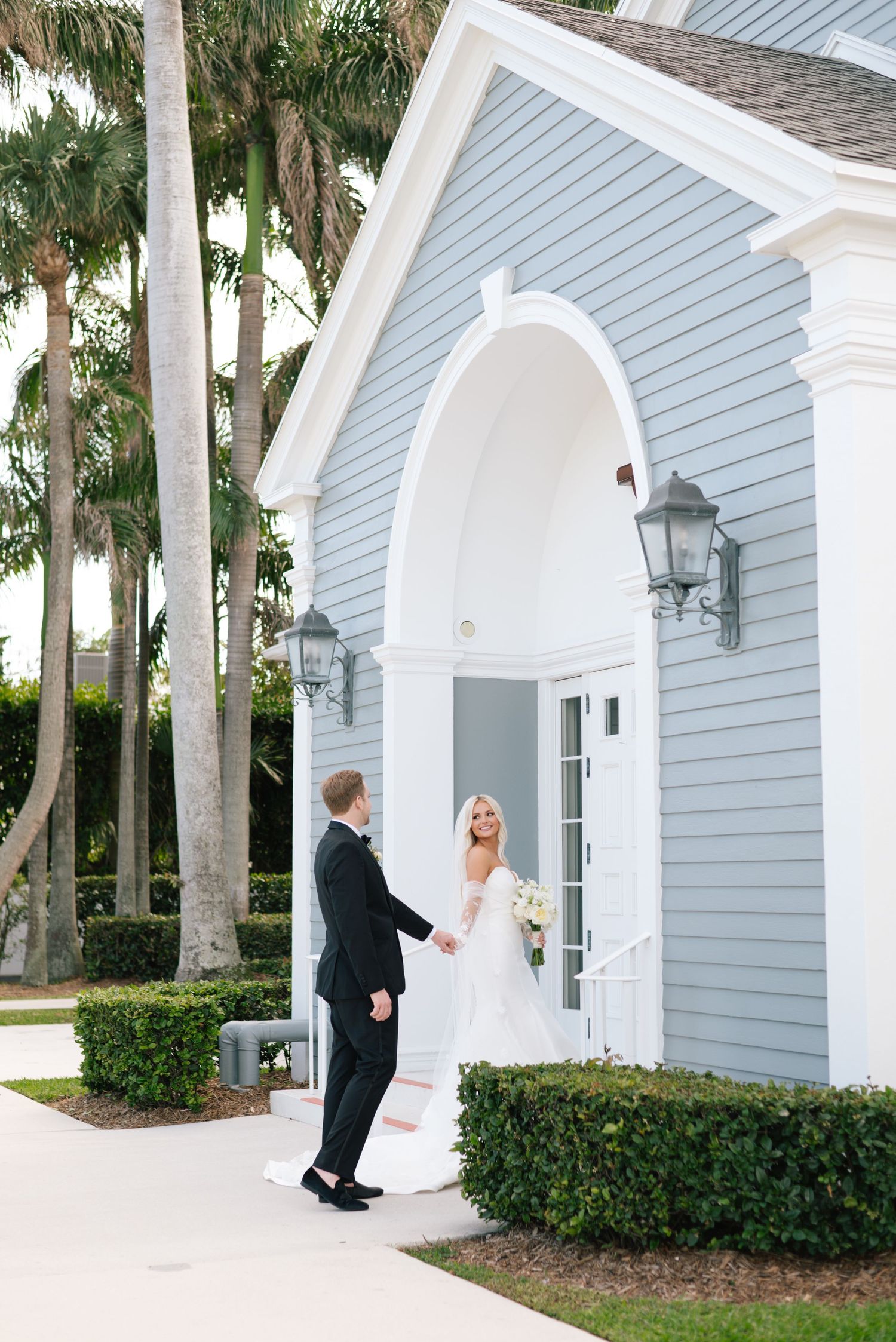 Wedding couple poses outside a light blue chapel with white columns and lanterns.