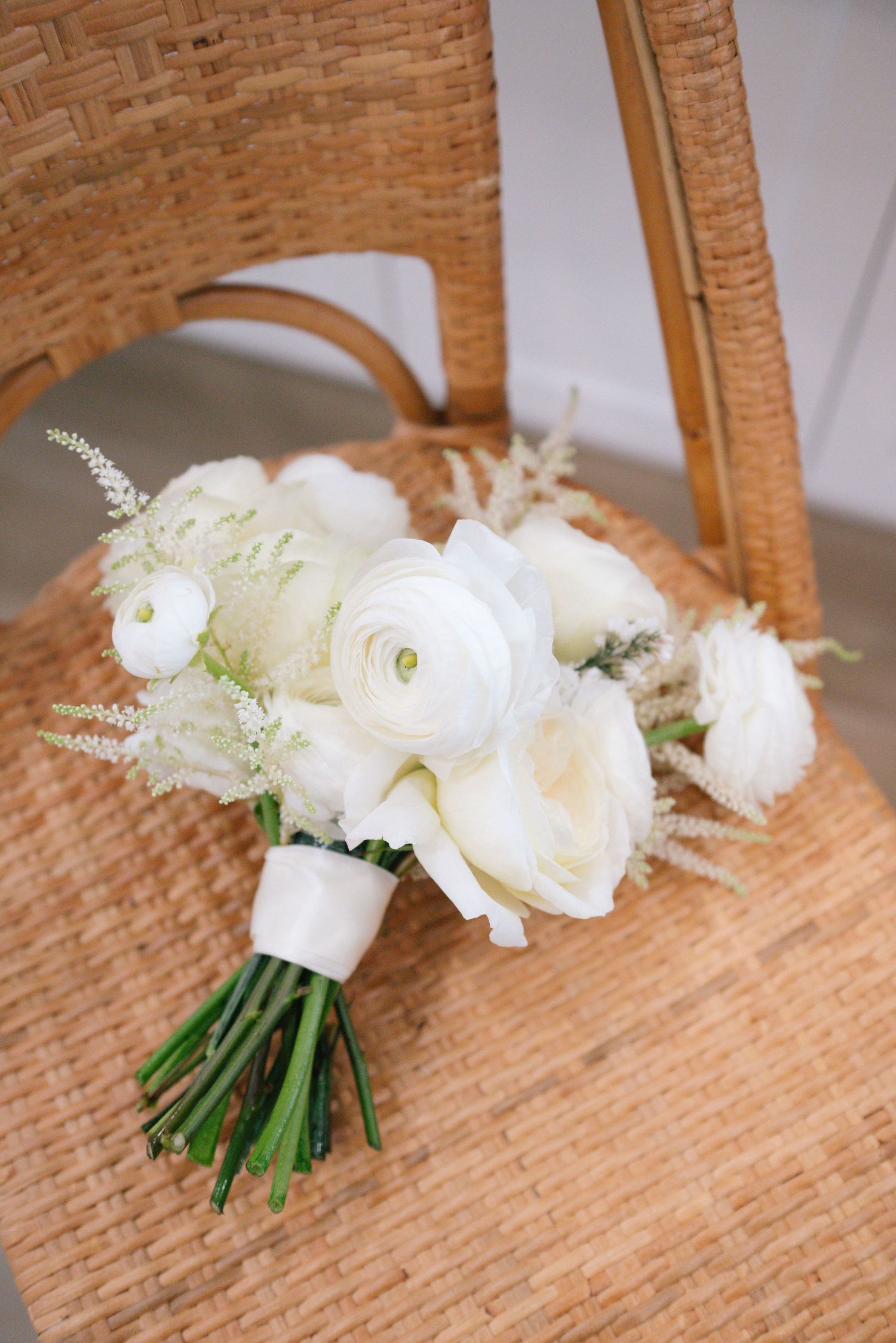 Elegant white bridal bouquet with ranunculus flowers and delicate greenery rests on a woven rattan chair.