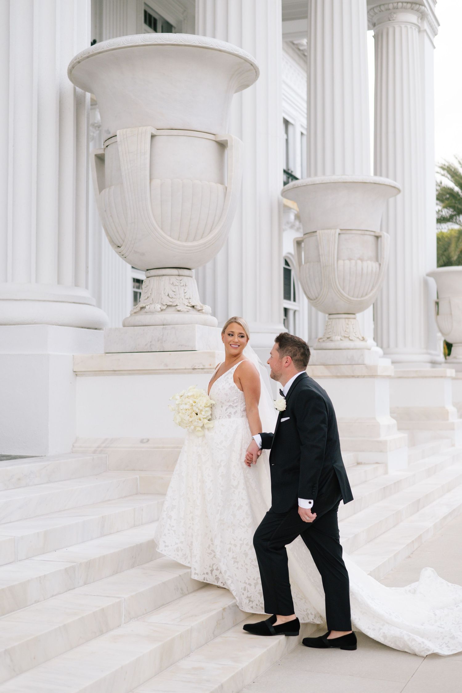 Bride and groom walk up white marble steps flanked by large decorative urns and elegant columns at a wedding venue.