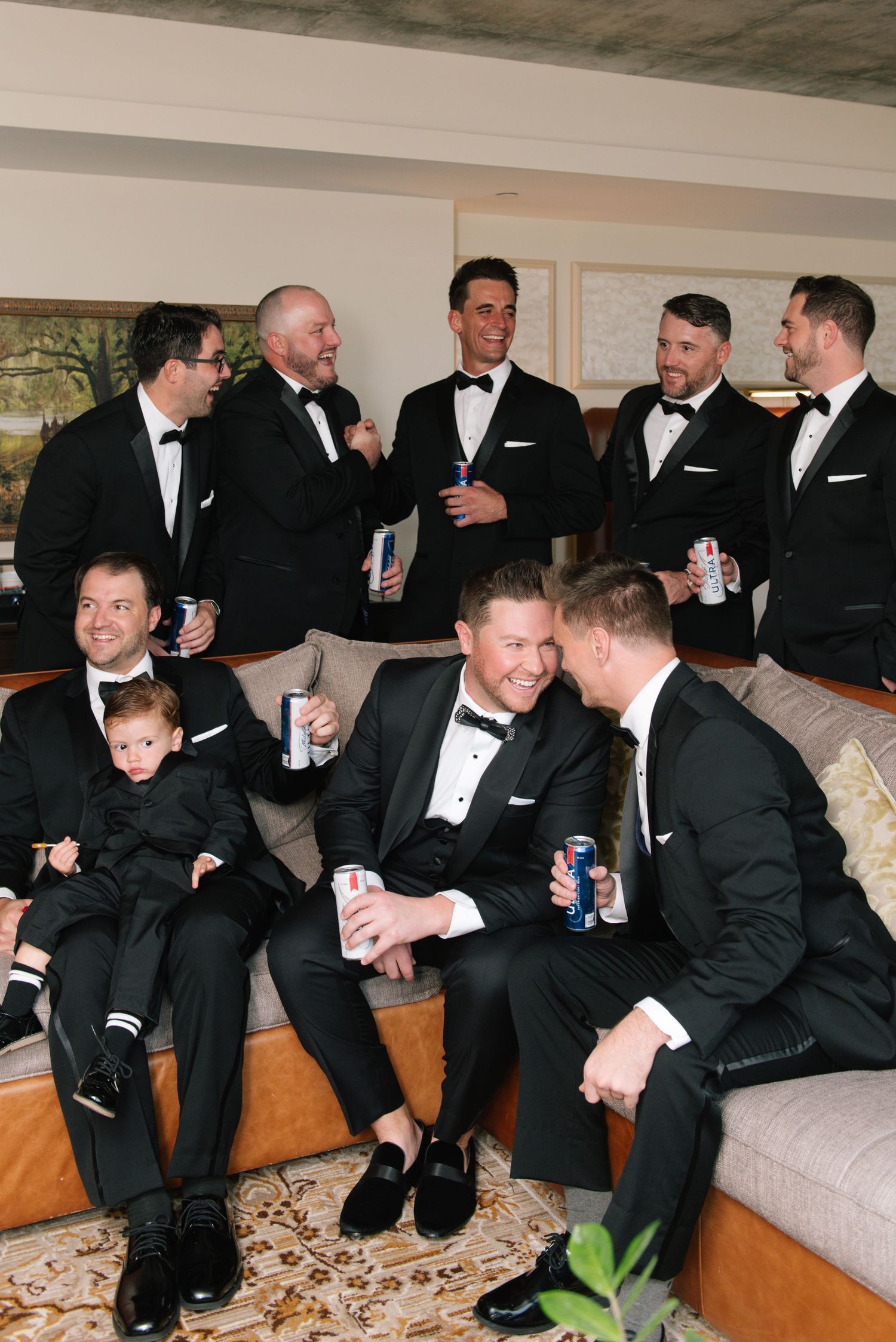 Groomsmen in black tuxedos relaxing with drinks before a wedding ceremony, including a young child in matching attire.